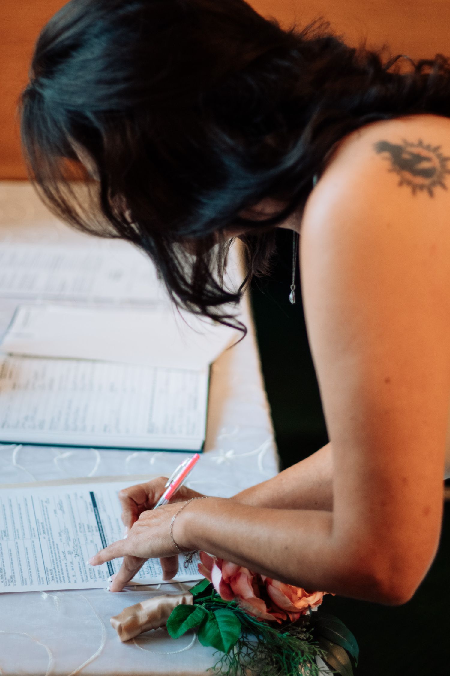 Bride with shoulder tattoo signs wedding documents with a red pen at a decorated table.