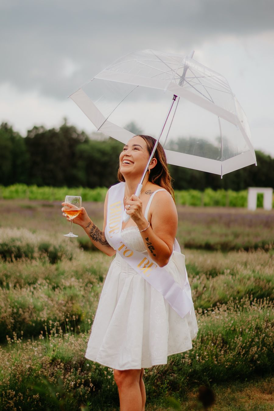 Smiling bride-to-be in white dress holding clear umbrella and wine glass in lavender field.