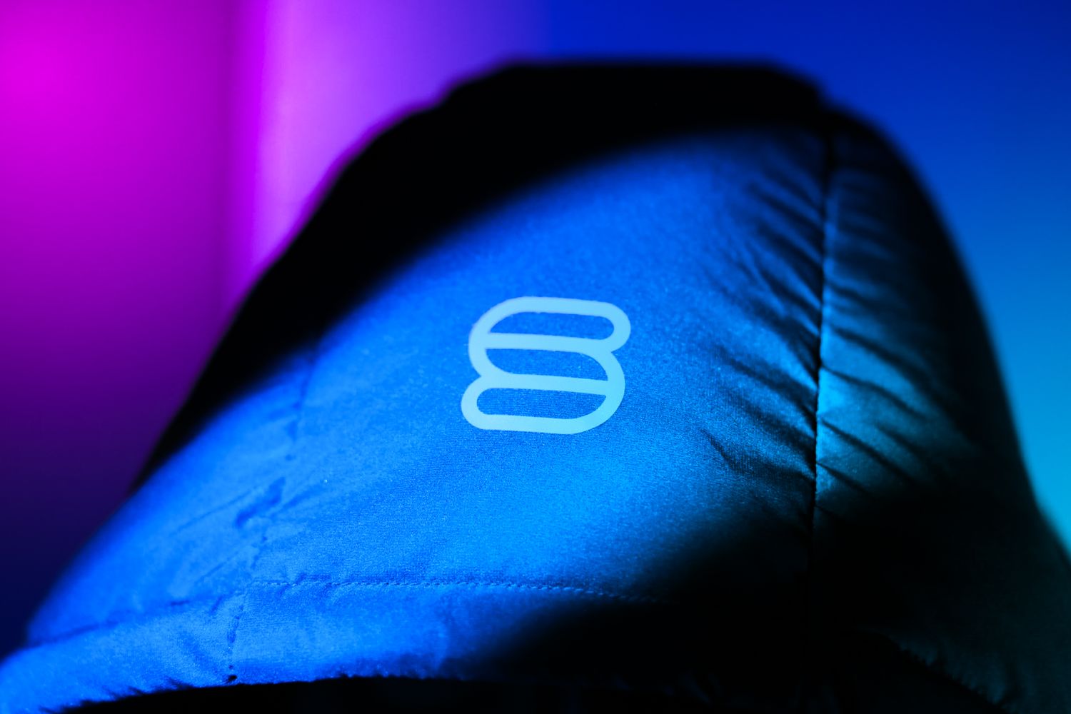 Close-up of quilted jacket shoulder featuring glowing brand logo under blue and pink neon lights.