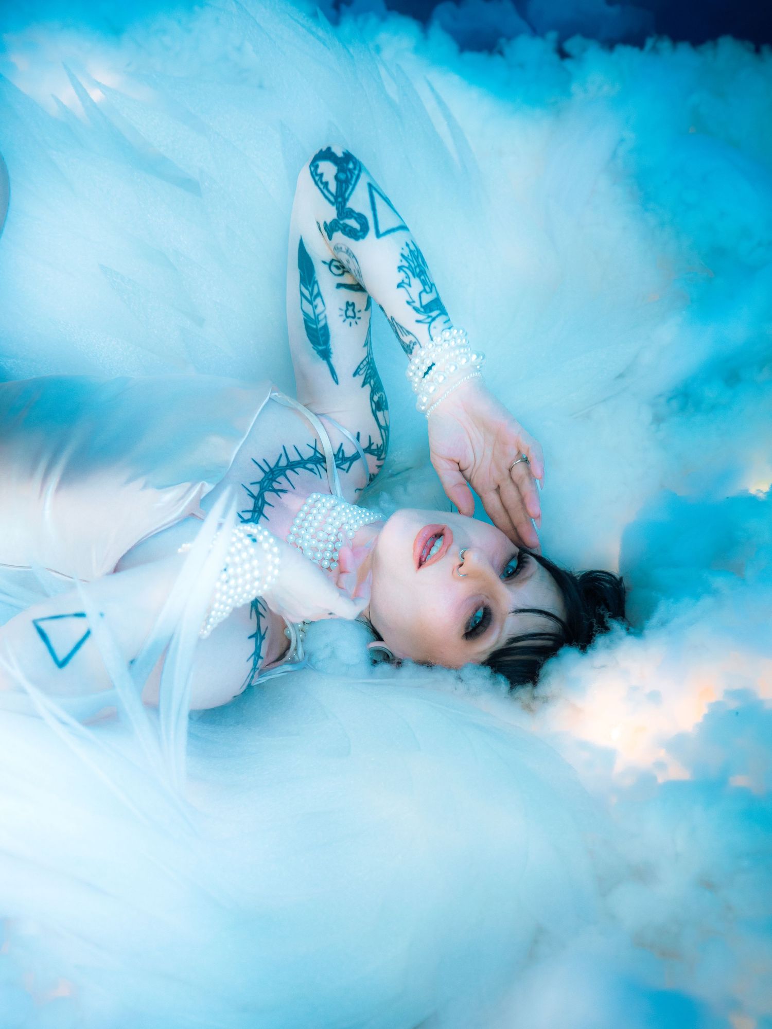 A tattooed woman in white tulle dress lies among blue clouds with pearl choker and dramatic makeup.