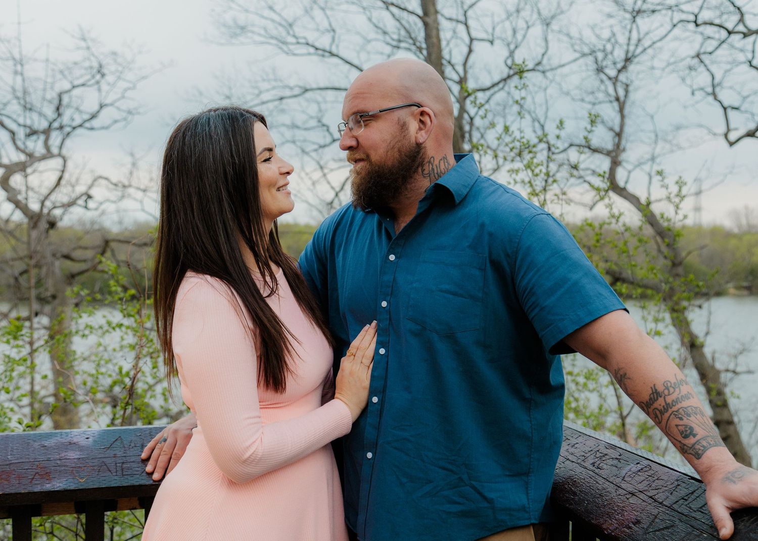 Couple smiling at each other on a wooden deck surrounded by trees and a lake in a romantic outdoor setting.