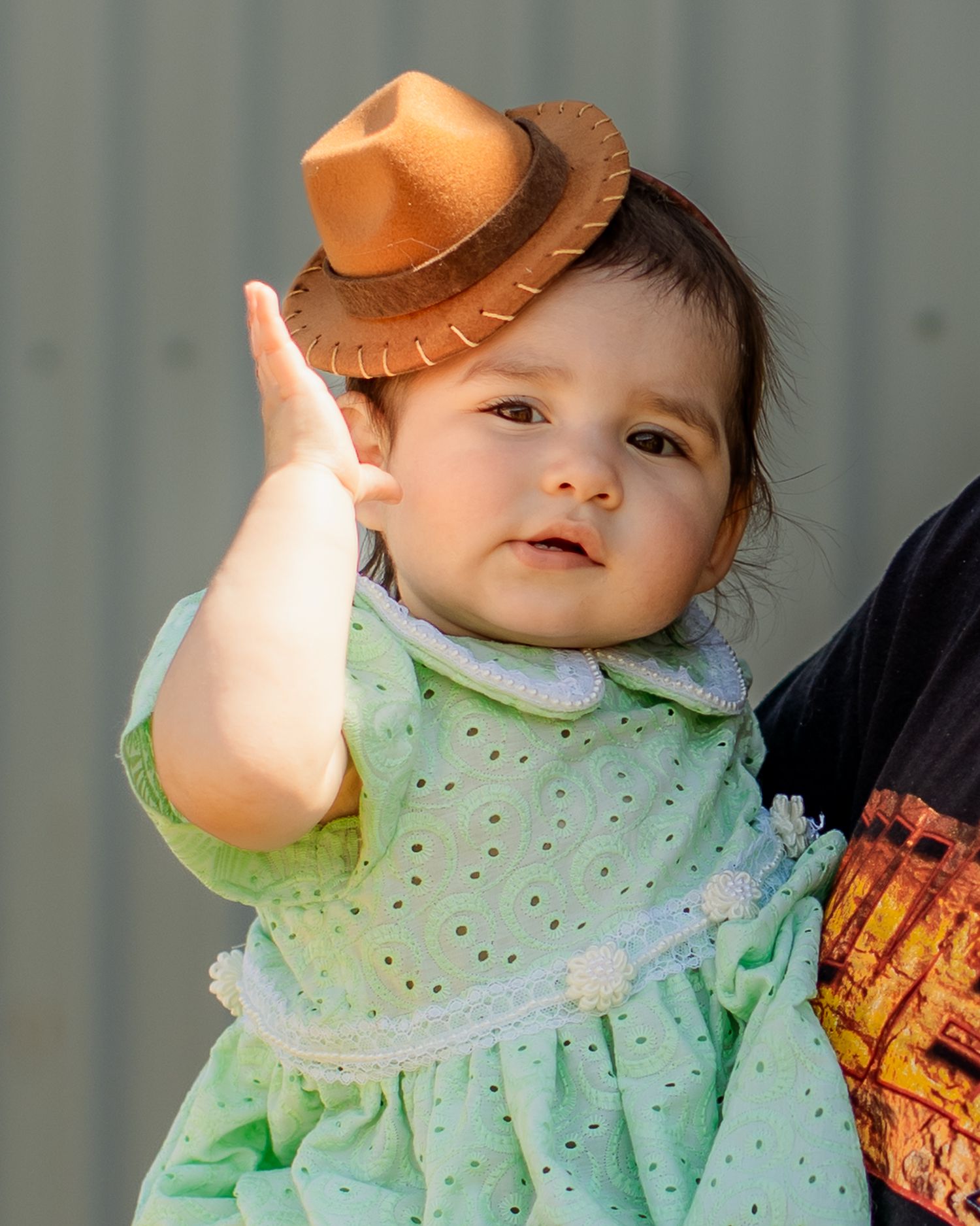 Adorable baby wearing a tiny cowboy hat and green eyelet dress, raising hand as if waving hello.
