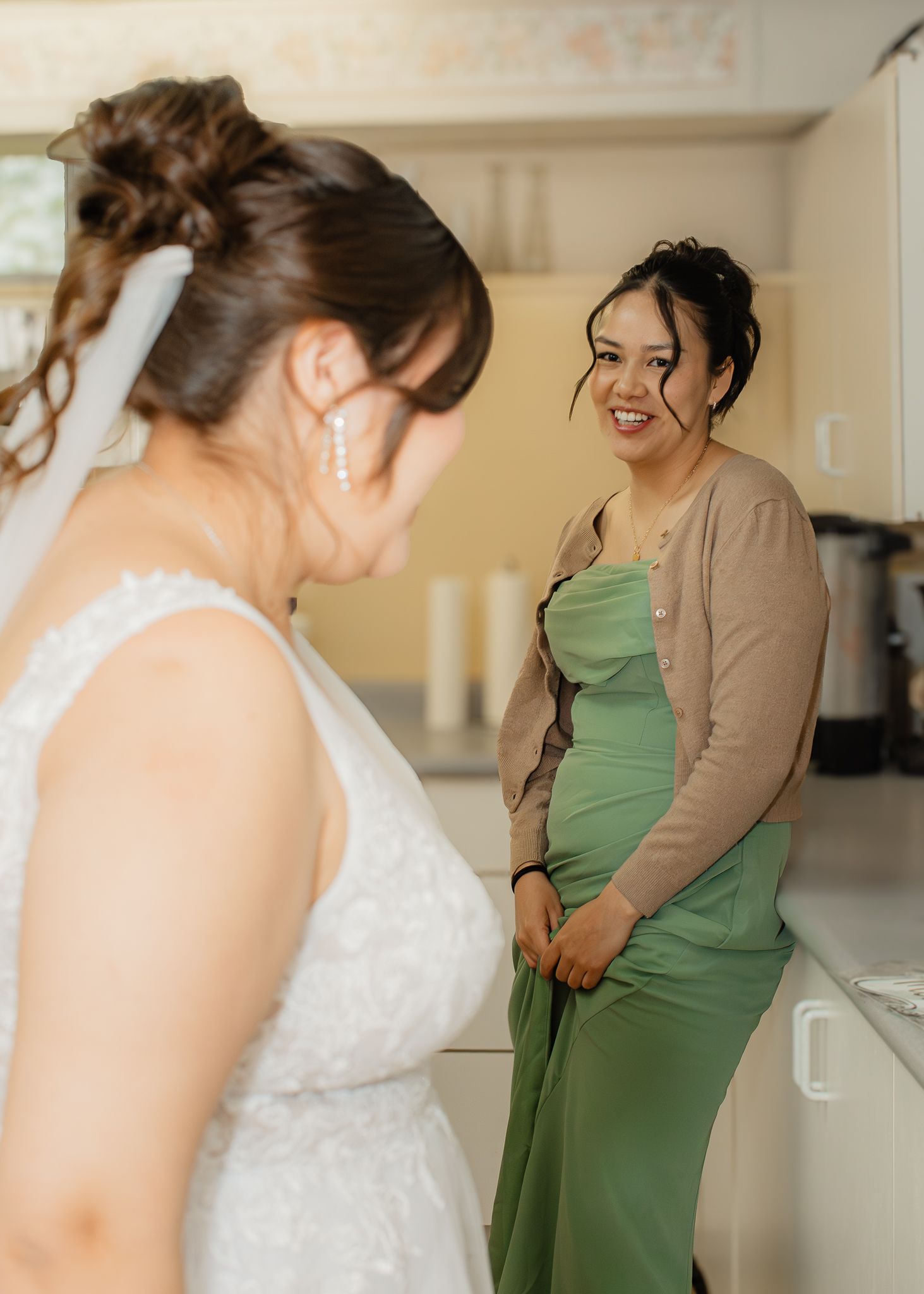 Bride in white lace gown smiles with bridesmaid in green dress and tan cardigan during wedding preparations.