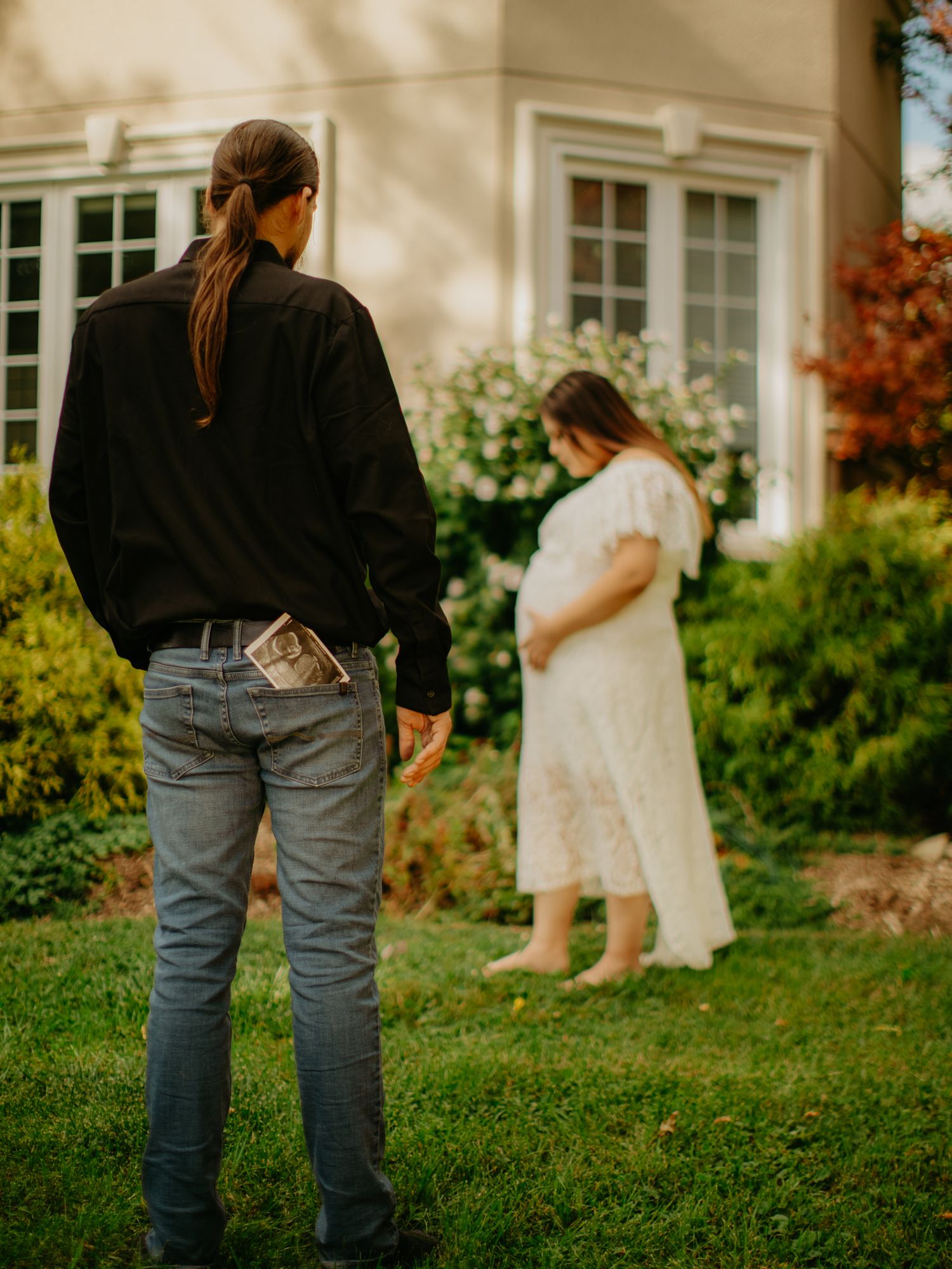 A couple poses in front of a house with white windows and fall foliage.