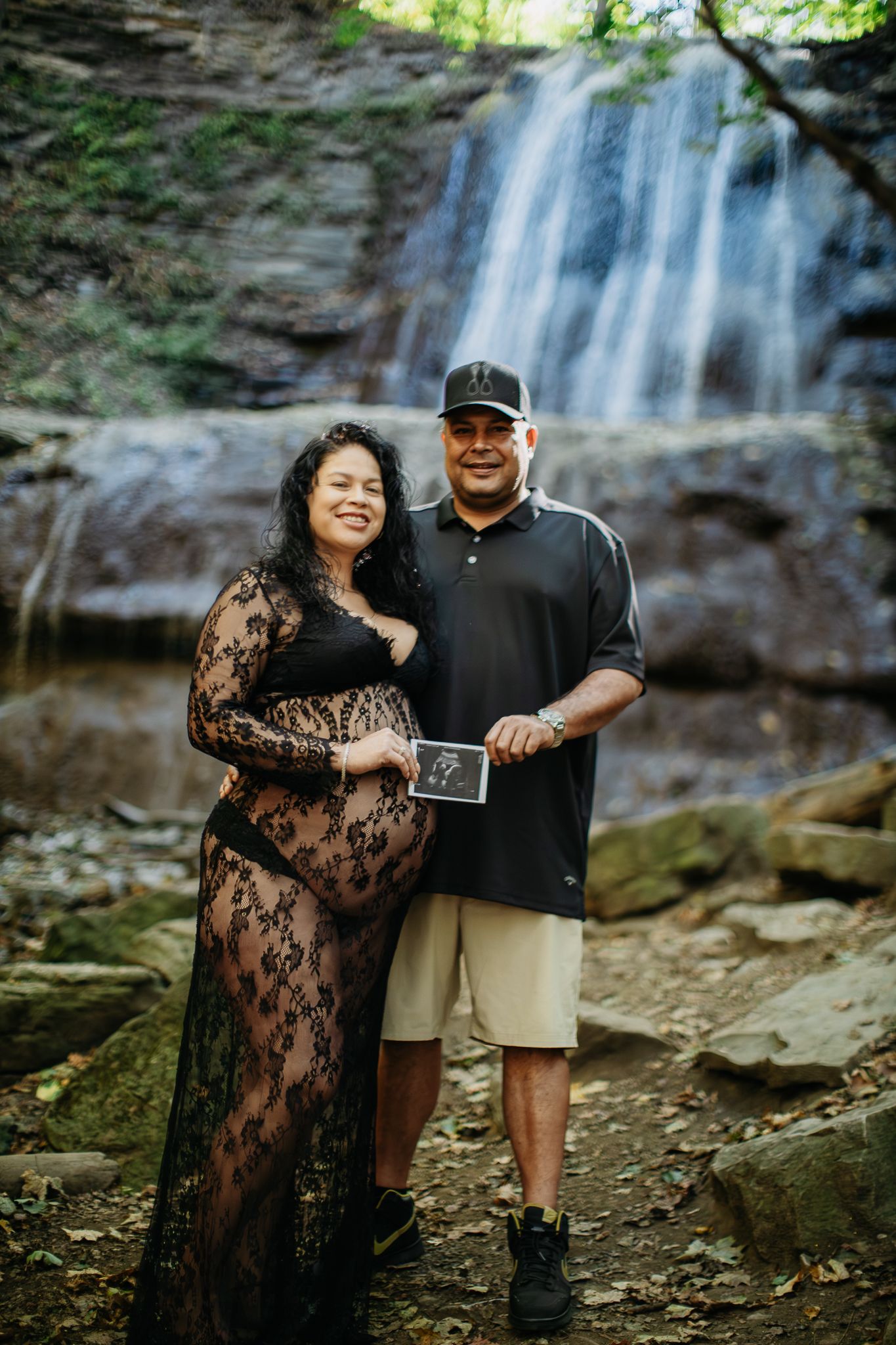 A couple poses together in front of a scenic waterfall in a romantic outdoor photoshoot.