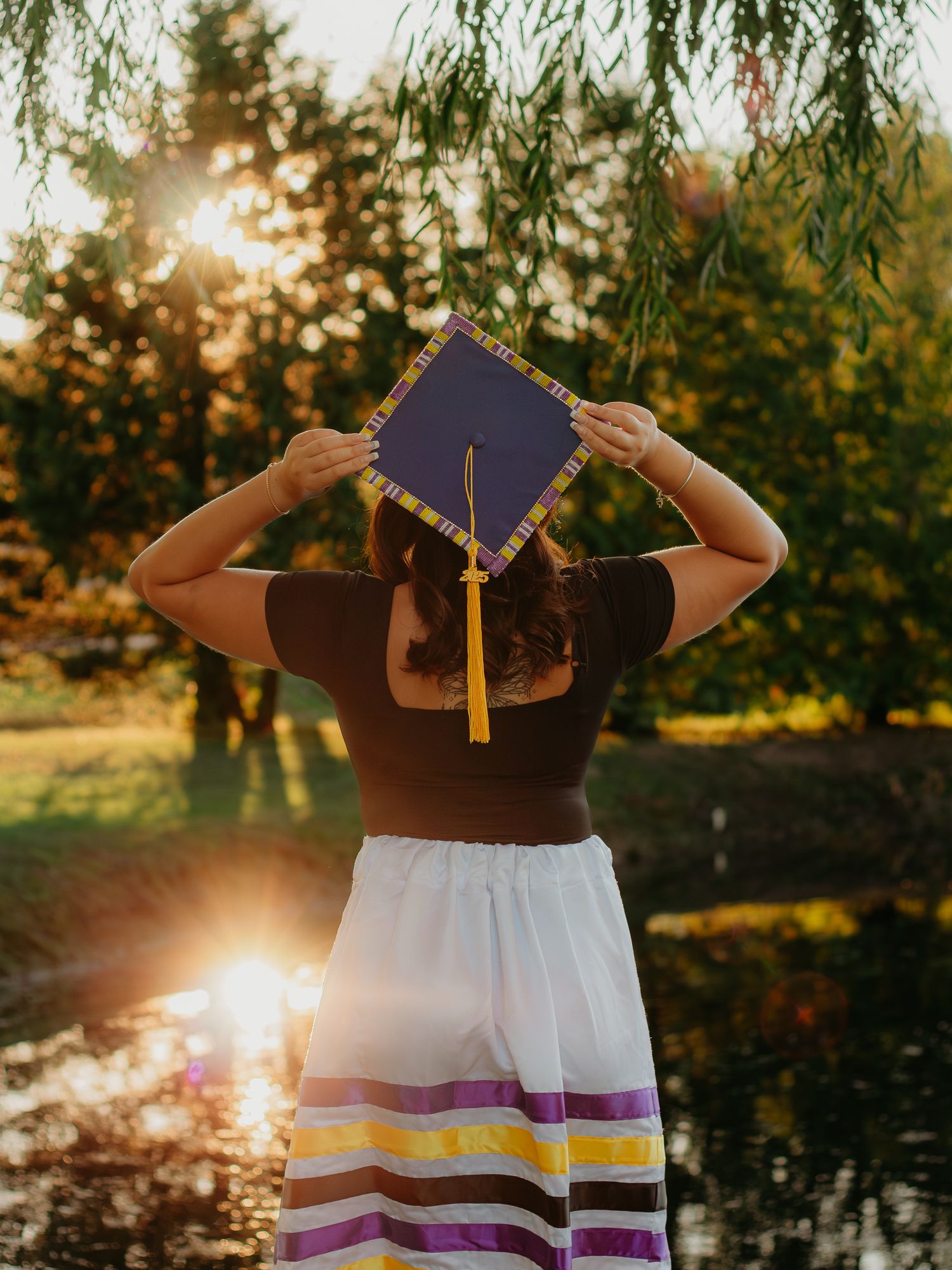 A graduate poses with their diploma against a sunset-lit pond backdrop.