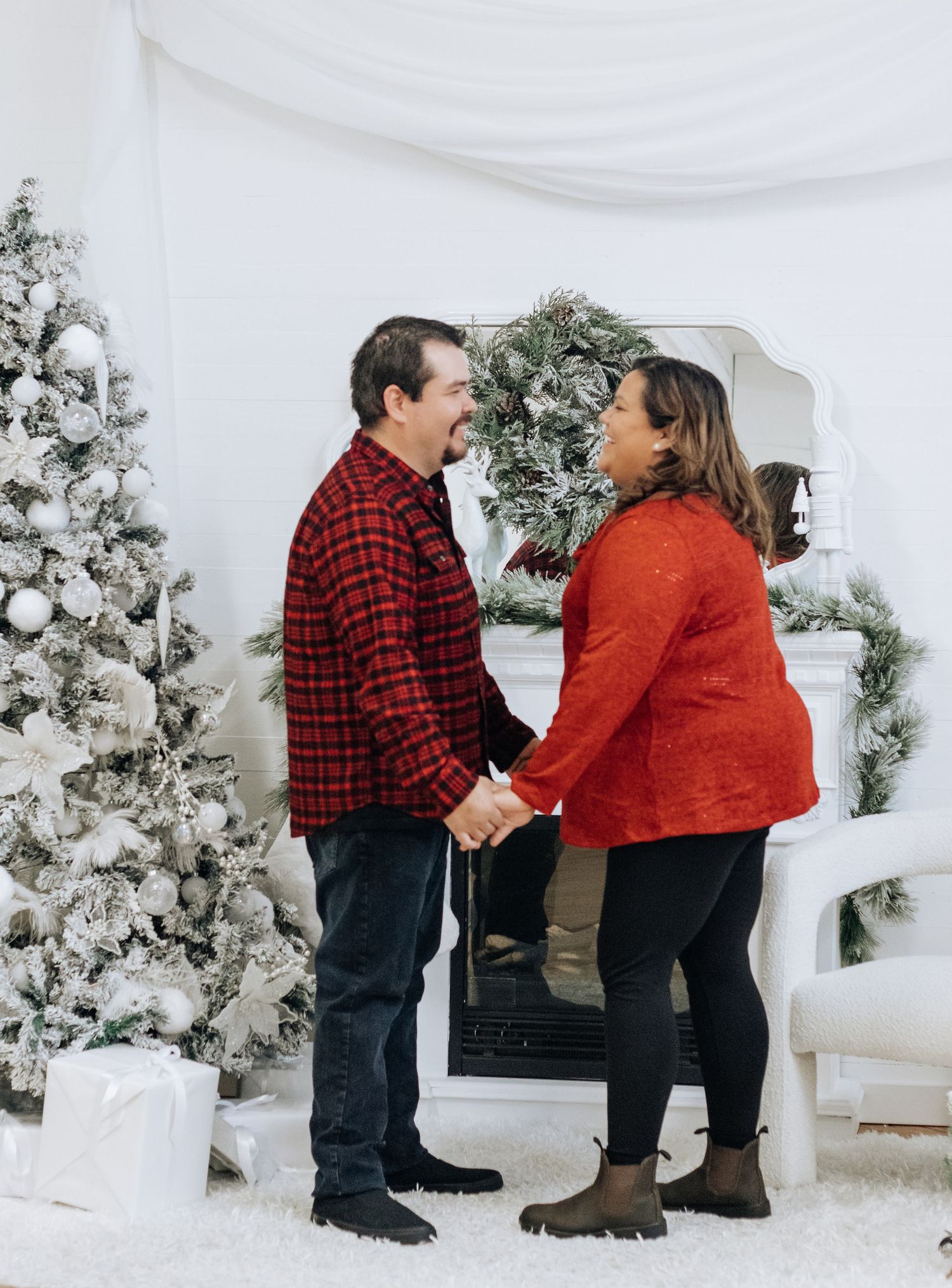 A couple holds hands in front of a decorated Christmas mantle with a flocked tree and white winter decor.