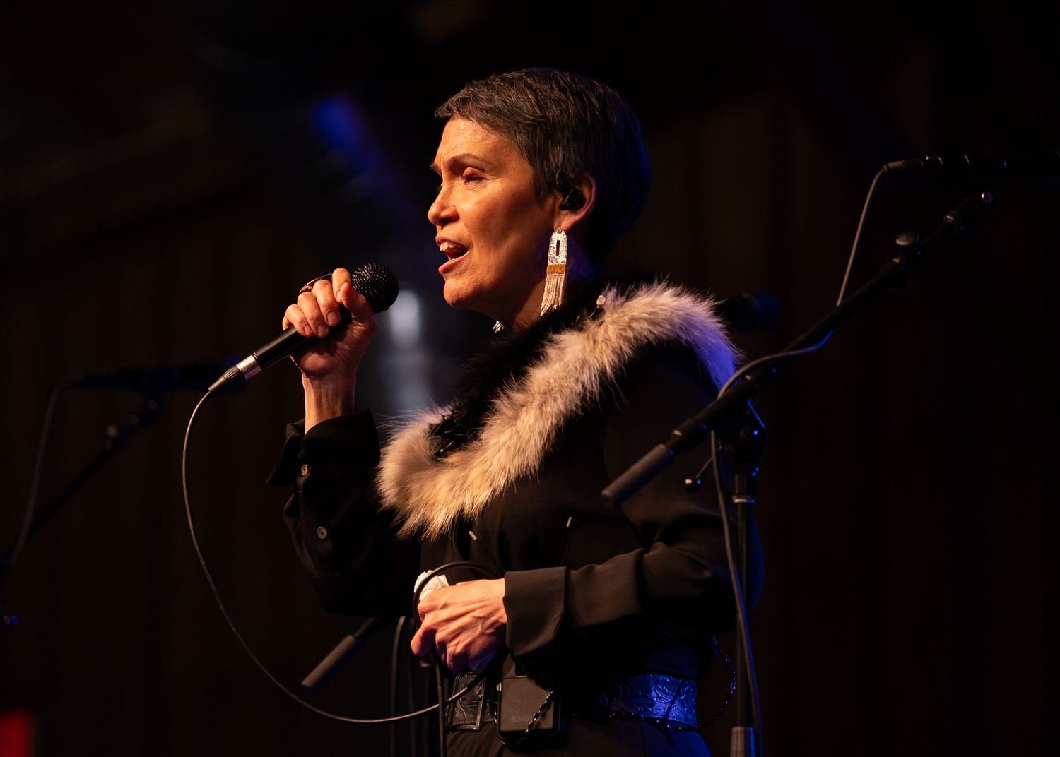 Woman singer performing on stage with microphone, wearing black jacket with fur collar under blue stage lighting.