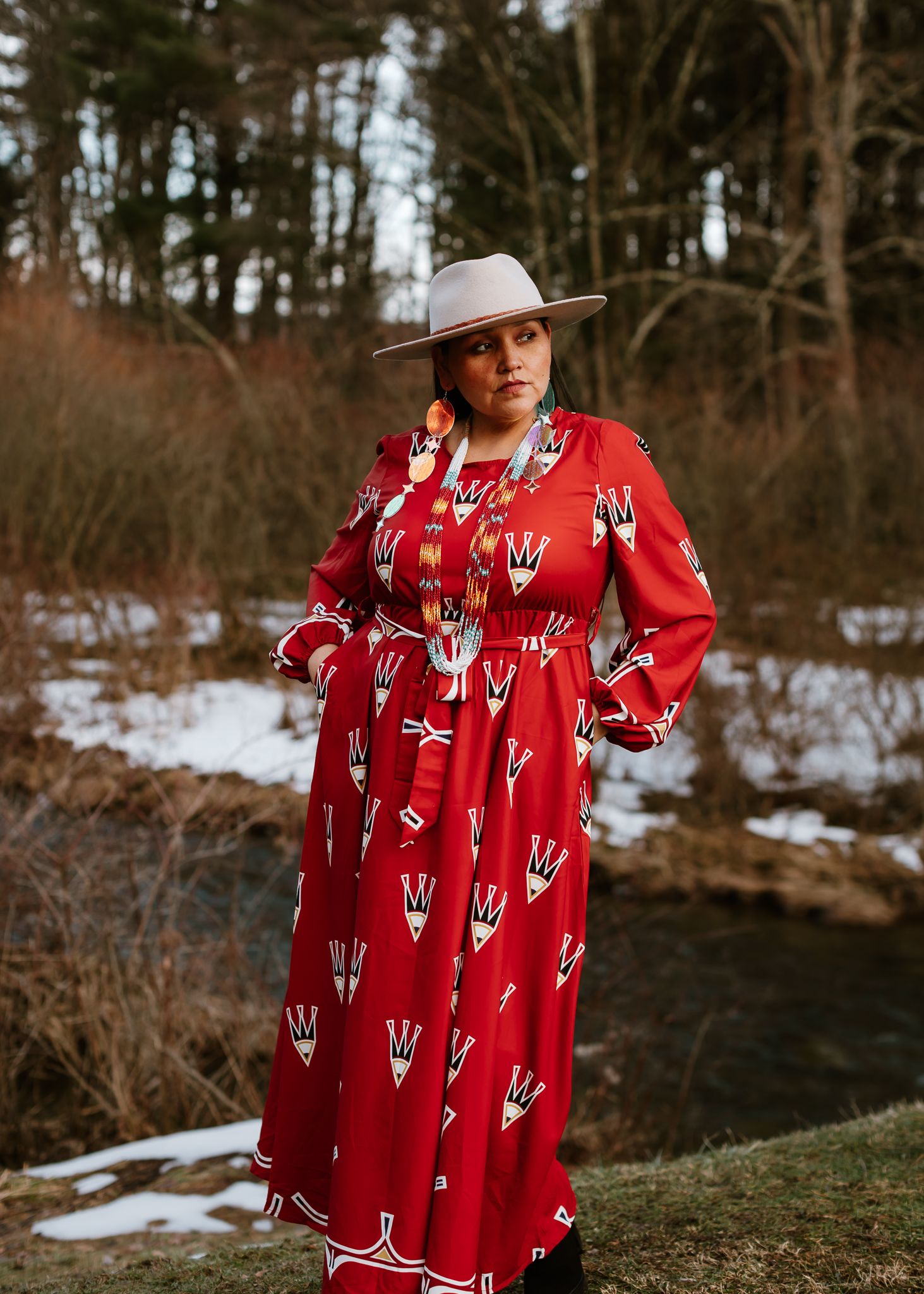 Woman in red patterned dress and wide-brim hat poses outdoors in snowy landscape with trees and stream.