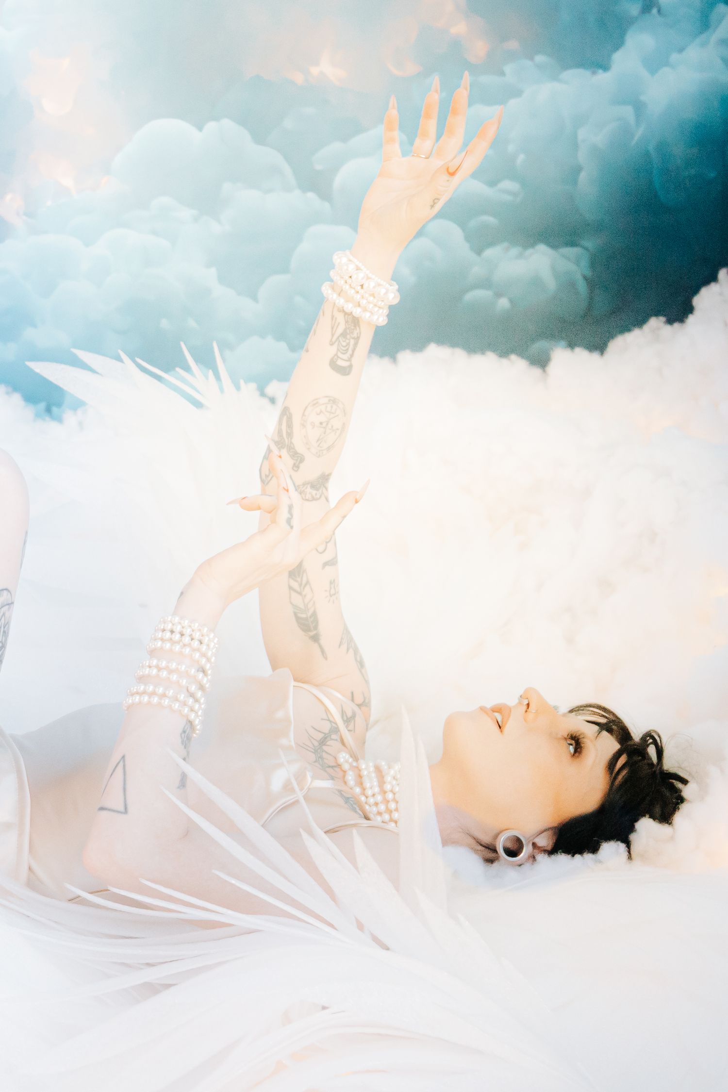 Tattooed woman in white angelic outfit reaching upward among clouds in a dreamy ethereal photo shoot.