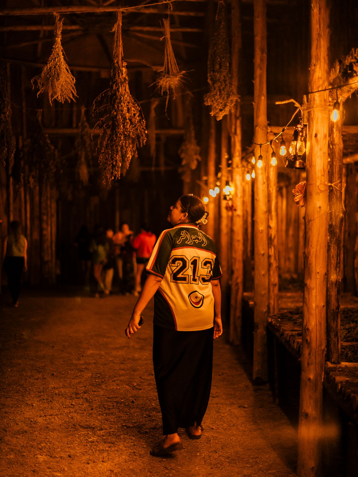Child walking through warmly lit rustic corridor with hanging dried herbs and string lights at night.
