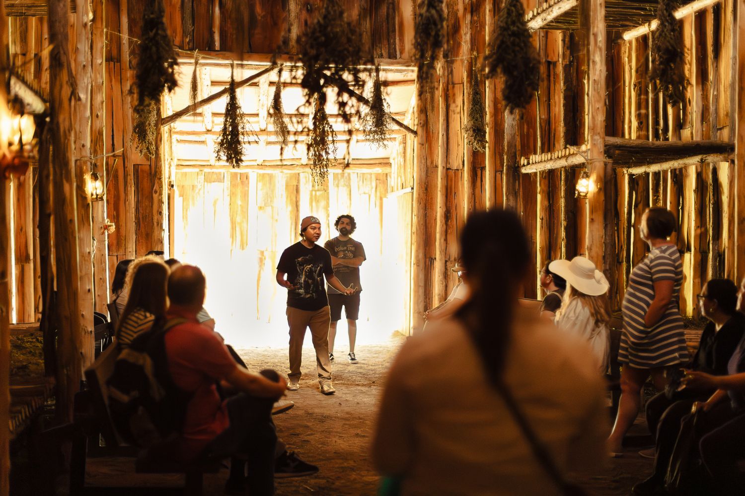People gather inside a rustic wooden barn filled with golden light and hanging dried herbs during an immersive art installation.