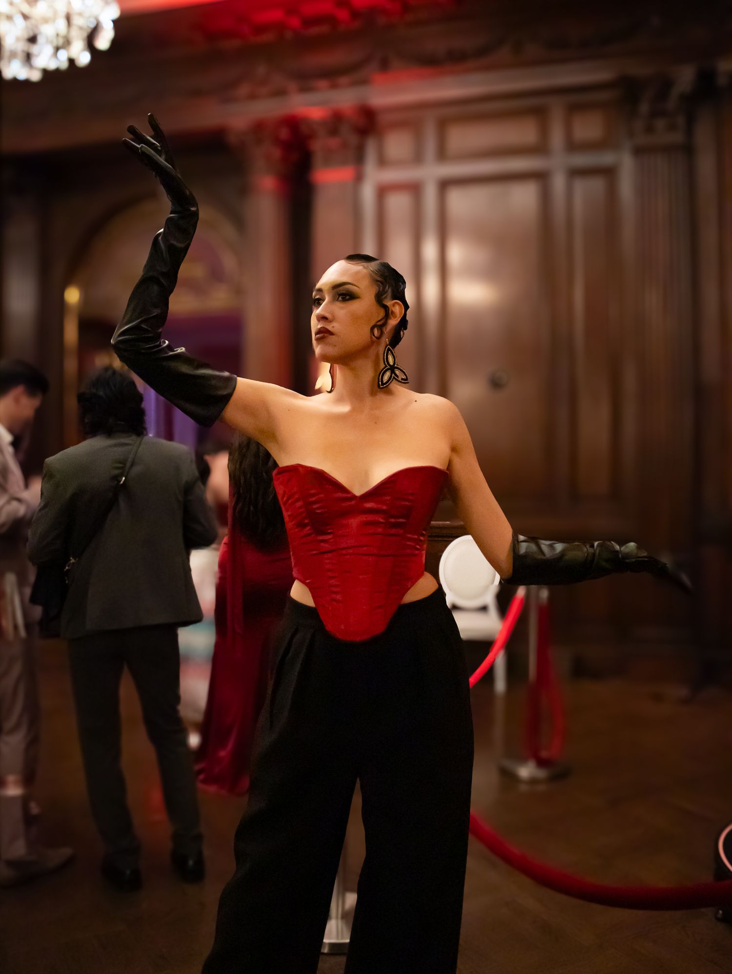 Woman in red velvet corset top, black pants and long gloves poses elegantly in an ornate wood-paneled ballroom.