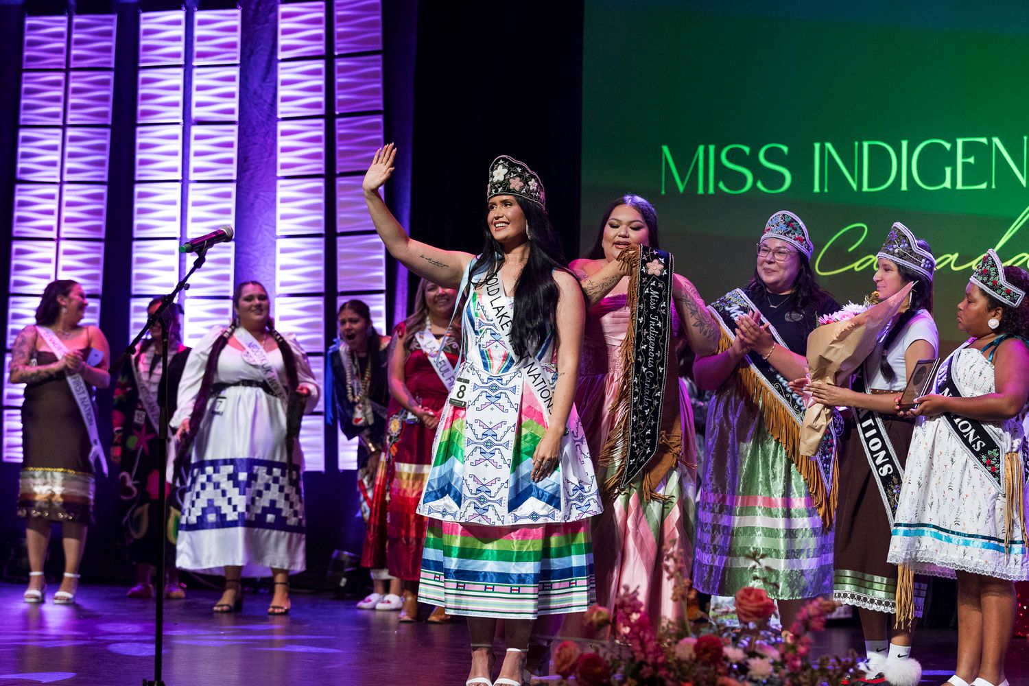 Miss Indigenous Canada winner celebrates on stage in colorful regalia surrounded by contestants.