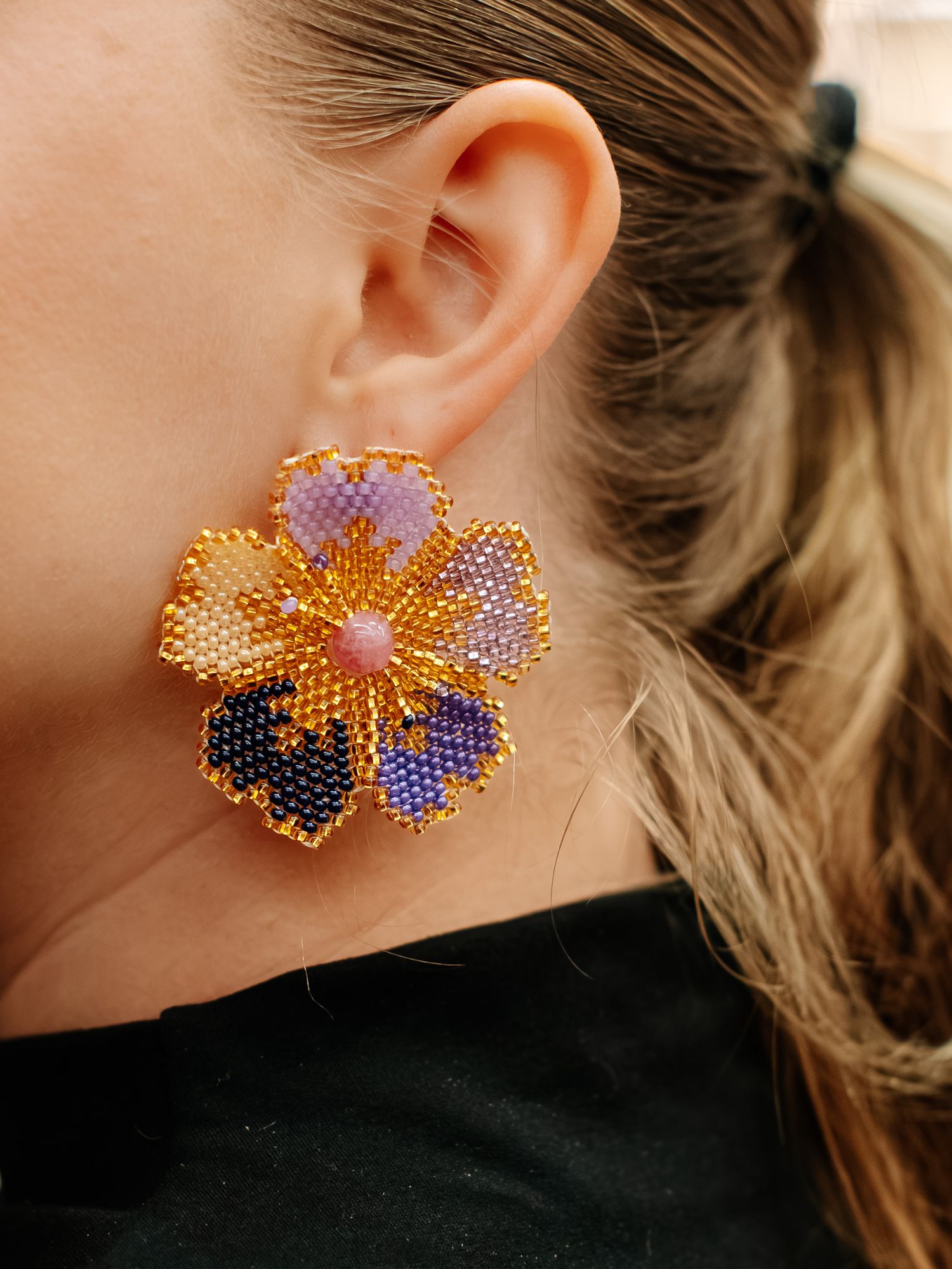 Woman wearing a large beaded flower earring in gold, purple, and blue tones with a pink center bead.