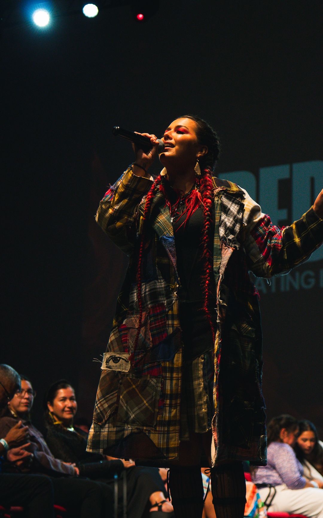 Singer performing on stage wearing a colorful patchwork coat while holding a microphone at a live event.