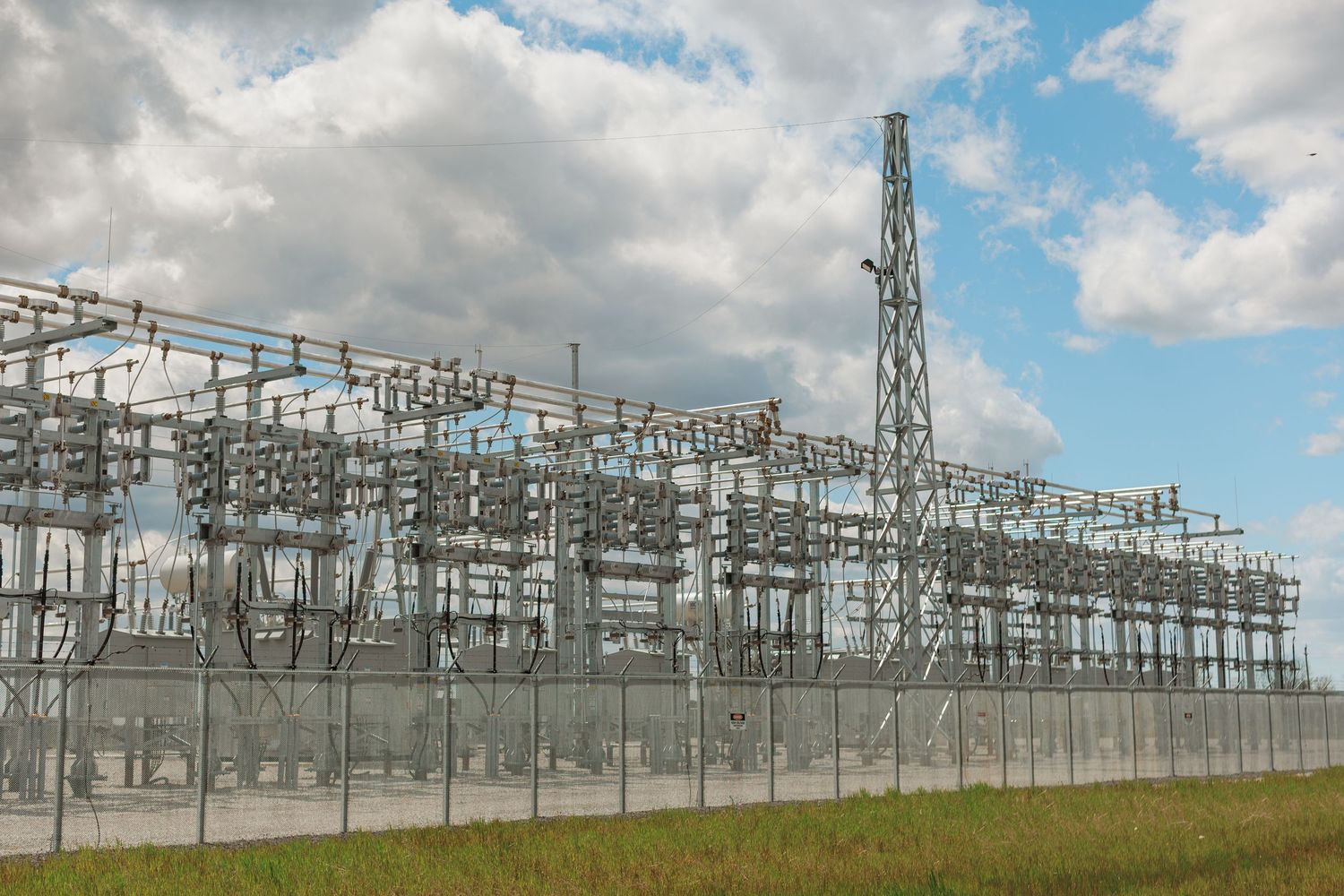 Large electrical substation with metal framework, transformers, and power lines behind a security fence under a cloudy sky.