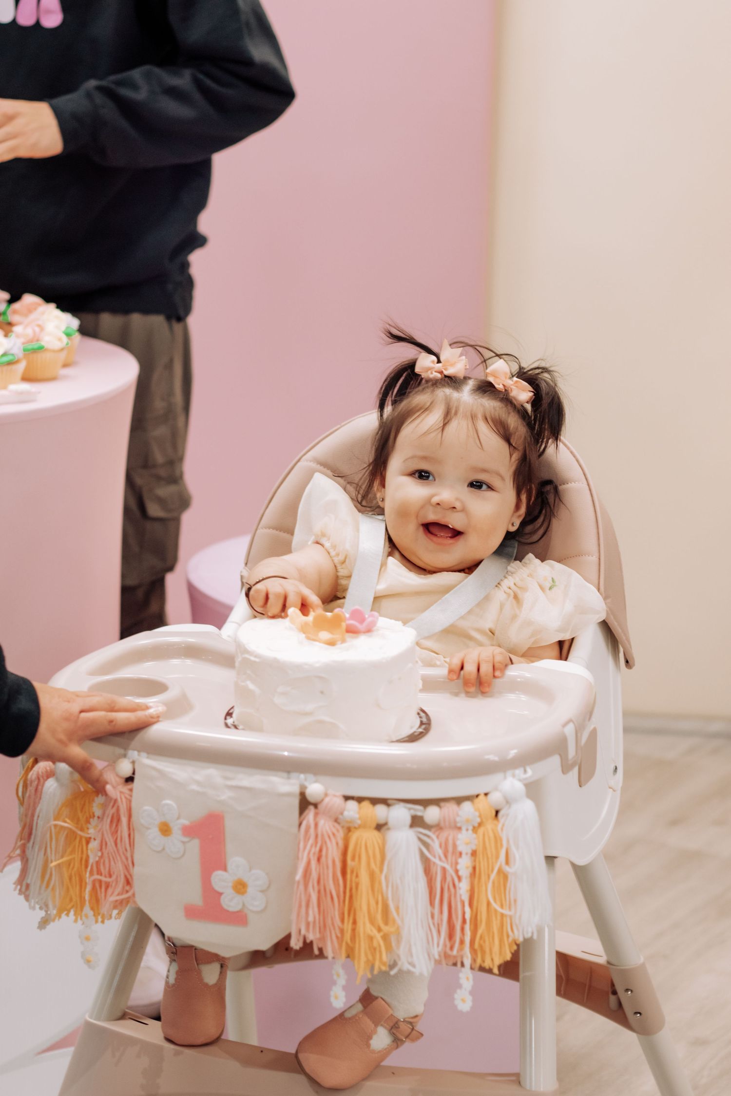 Smiling baby girl in decorated high chair celebrating first birthday with white cake and colorful tassel banner.
