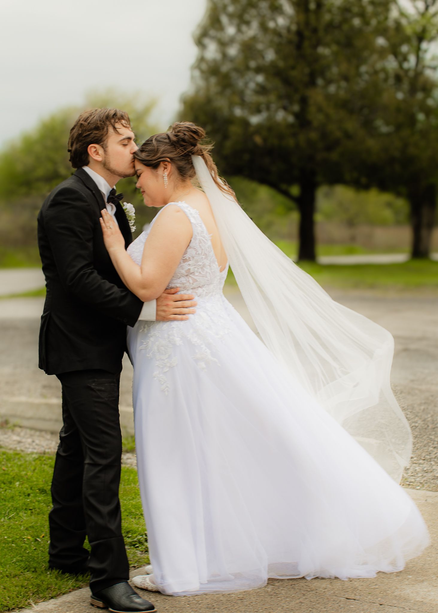 Bride and groom share romantic kiss outdoors with wedding veil flowing in the wind.