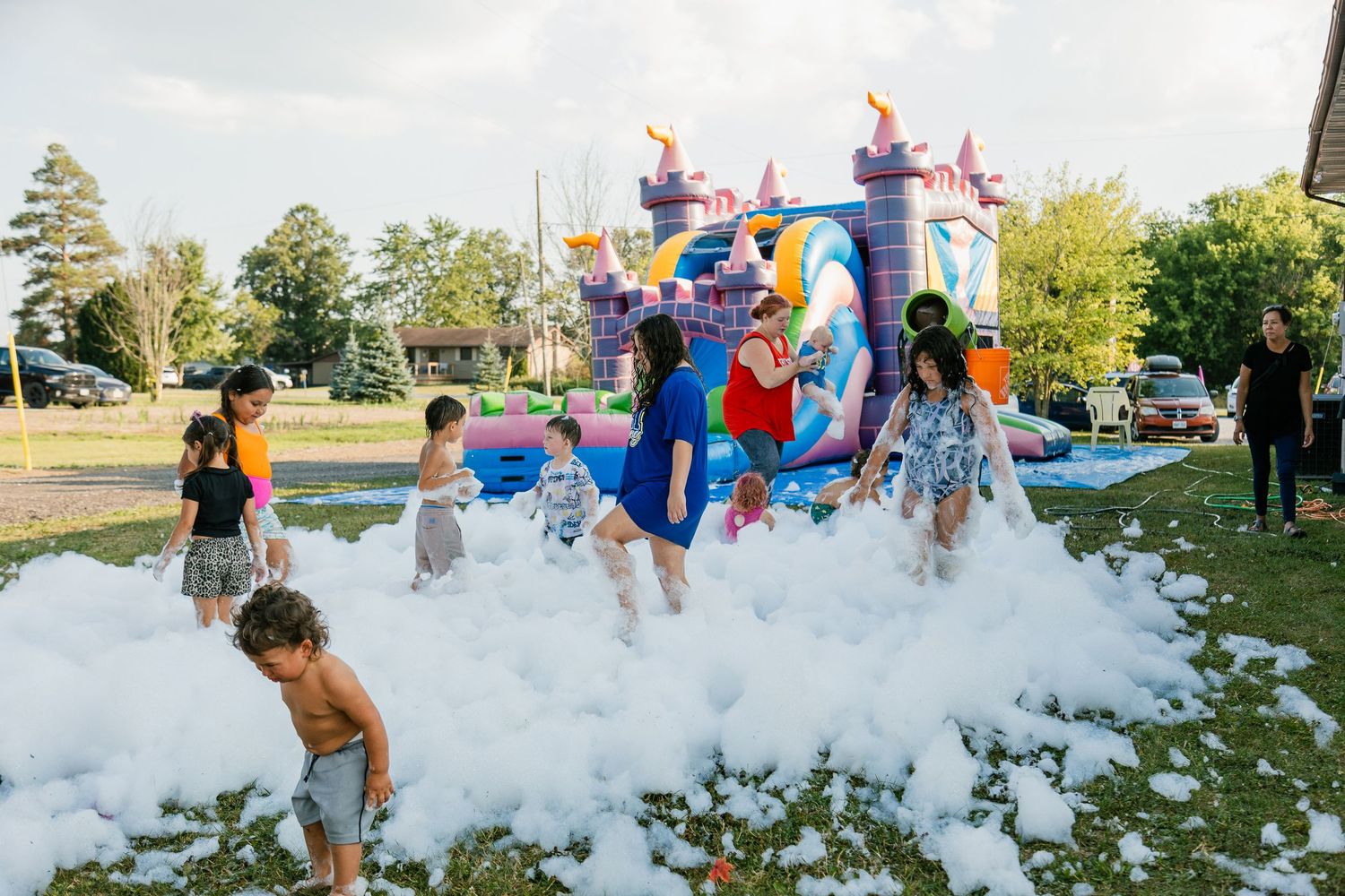 Kids playing in a foam party with a bouncy castle in the background at an outdoor event.