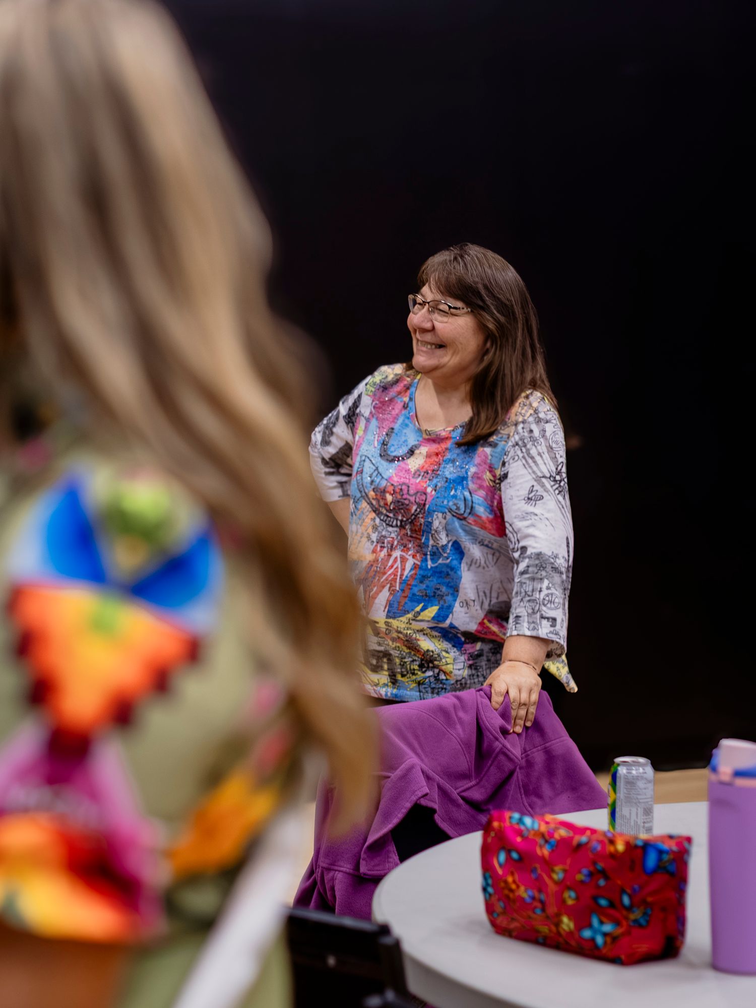 Woman in colorful floral top and purple skirt smiling during an indoor event with blurred foreground figure.