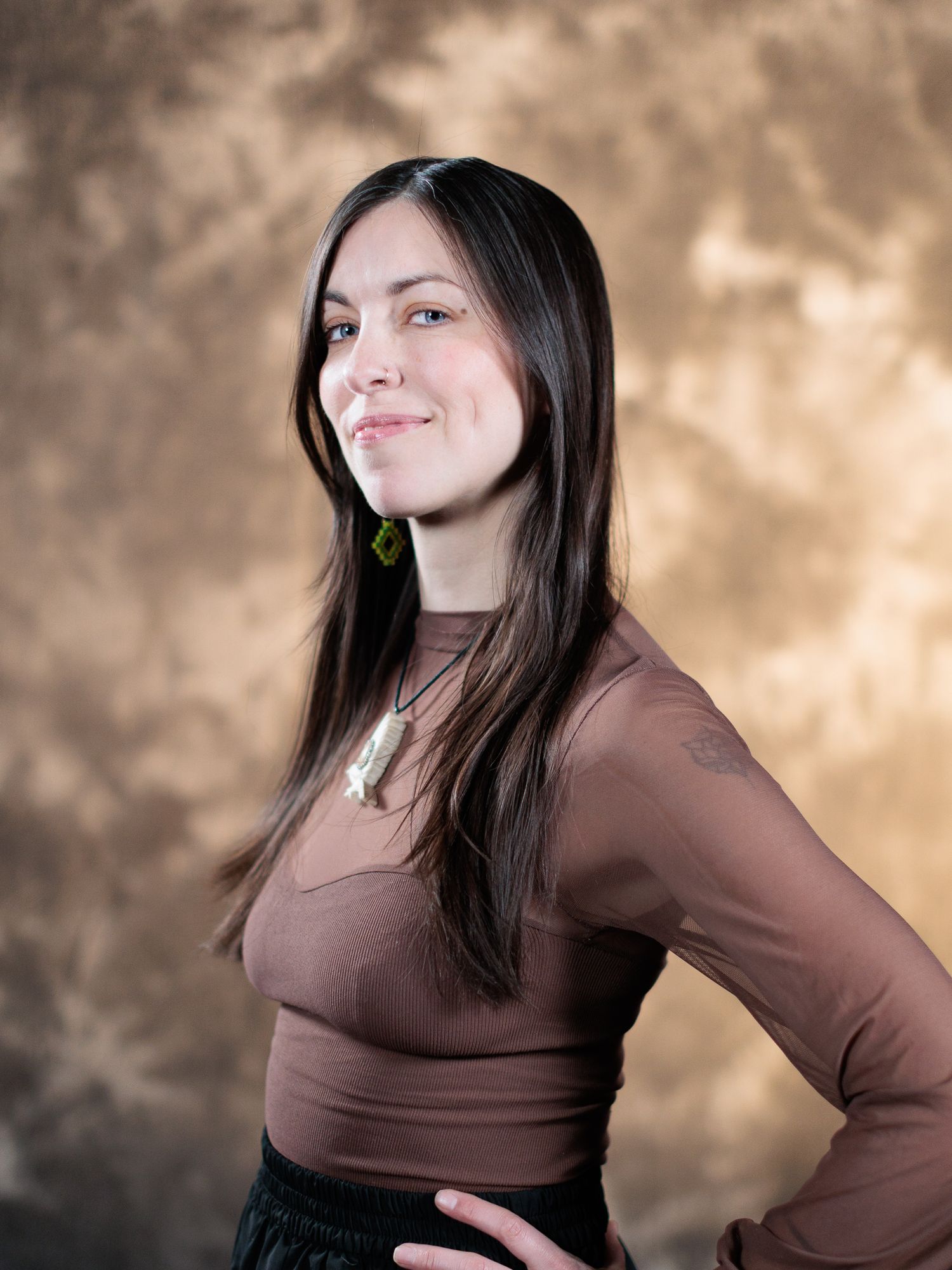 Young woman with long dark hair smiling, wearing a brown ribbed top and necklace, posed against a mottled brown backdrop.