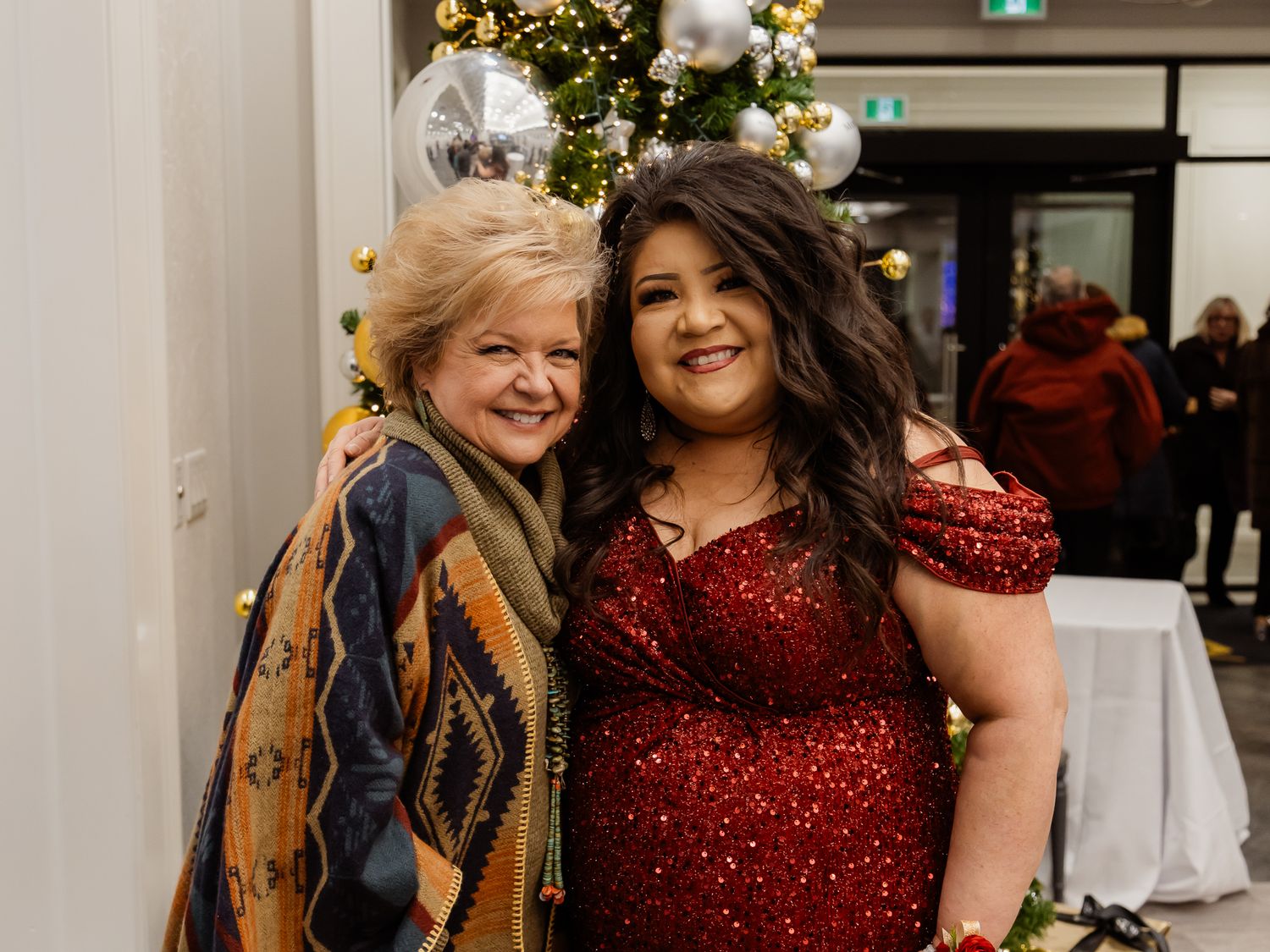 Two guests pose together at a holiday celebration with a decorated Christmas tree visible.