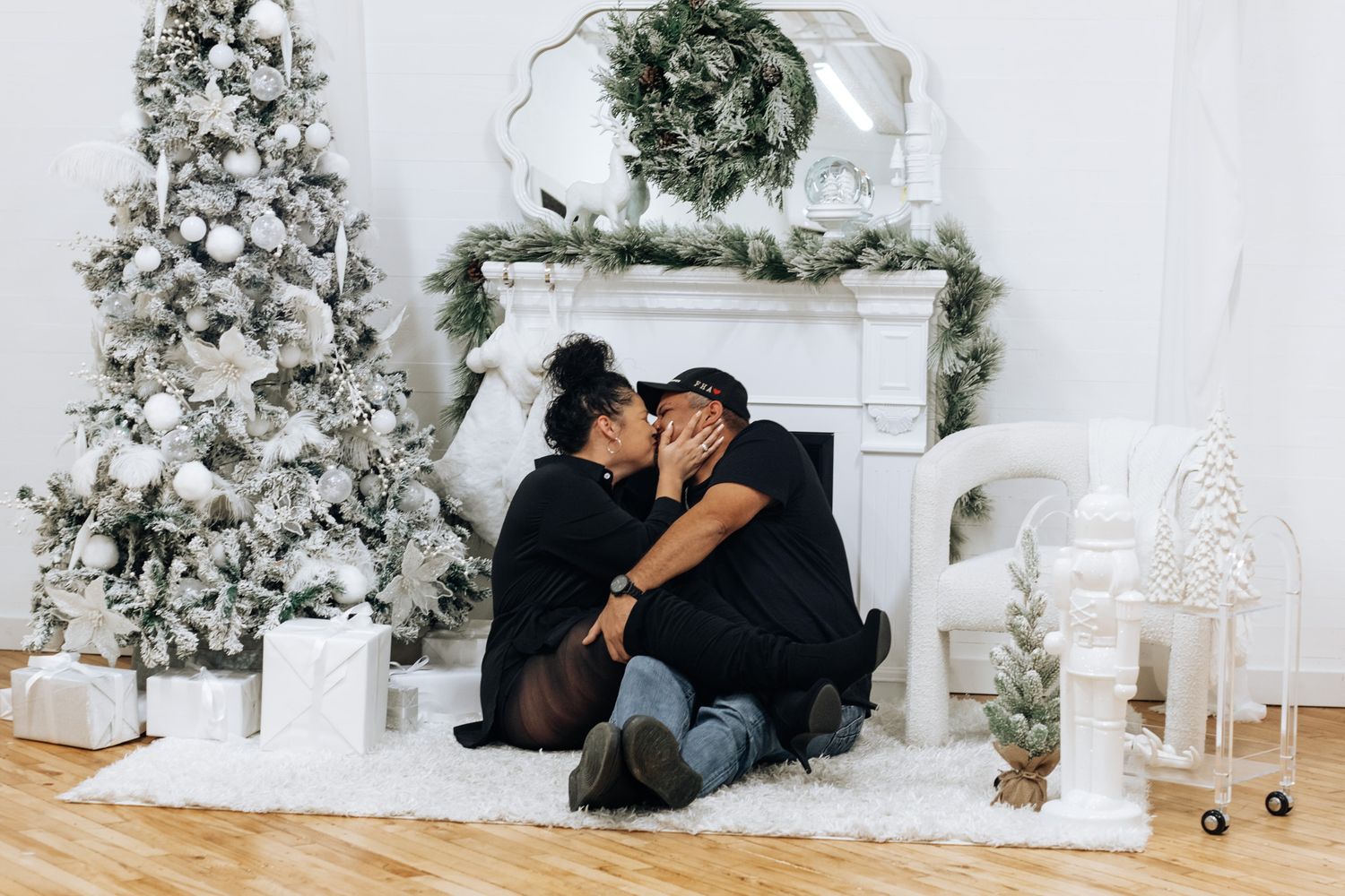 A couple embraces on the floor in front of a decorated Christmas fireplace and flocked tree.