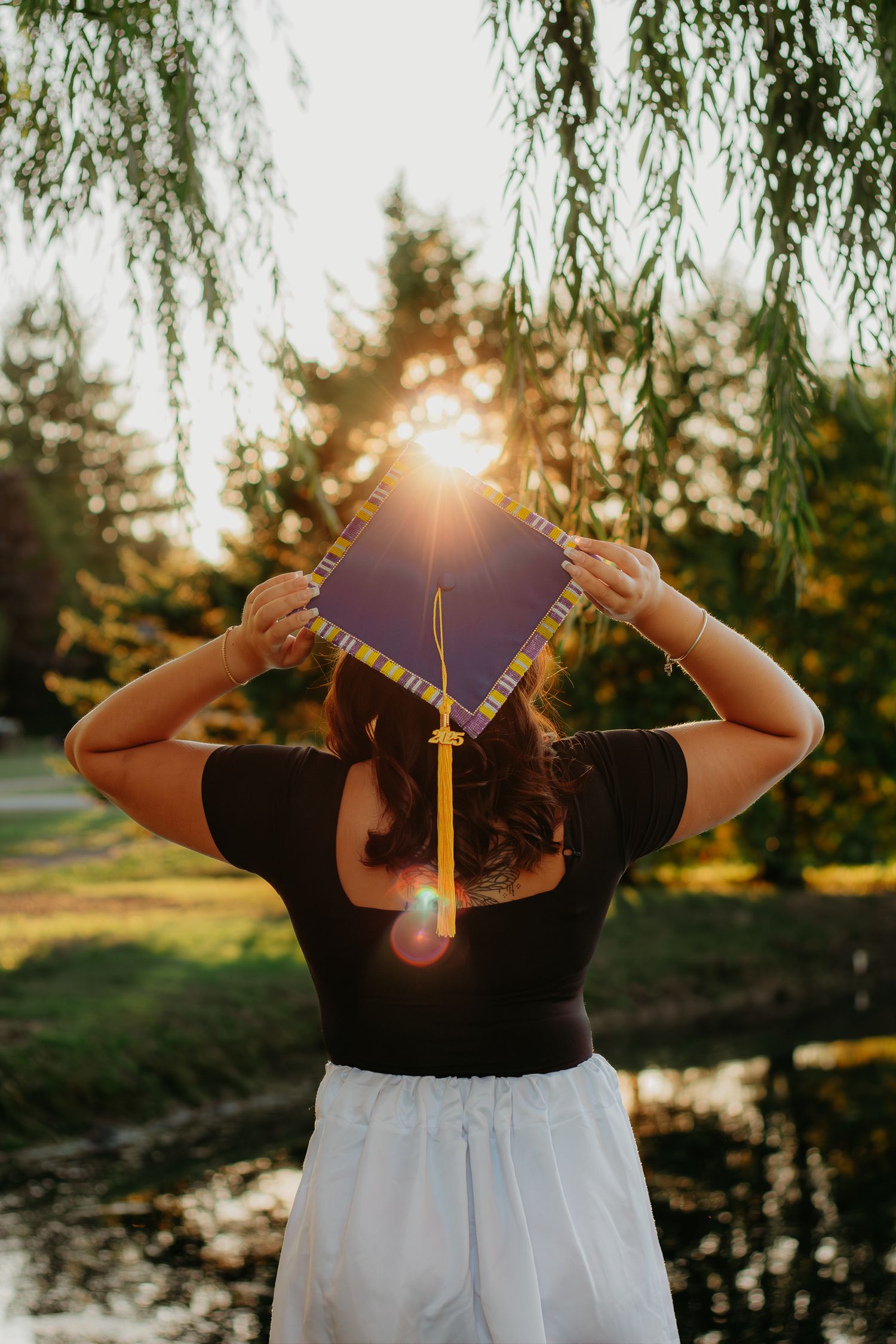 A graduate holds their cap while standing near a pond during golden hour.