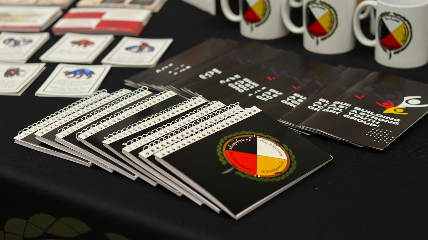 Row of printed materials and documents with indigenous medicine wheel logos displayed on dark tables.