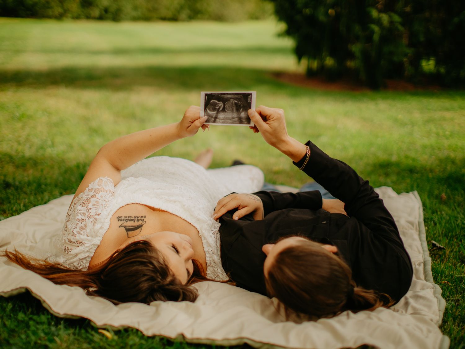 Two people relax on a white blanket outdoors while looking at a photograph together.