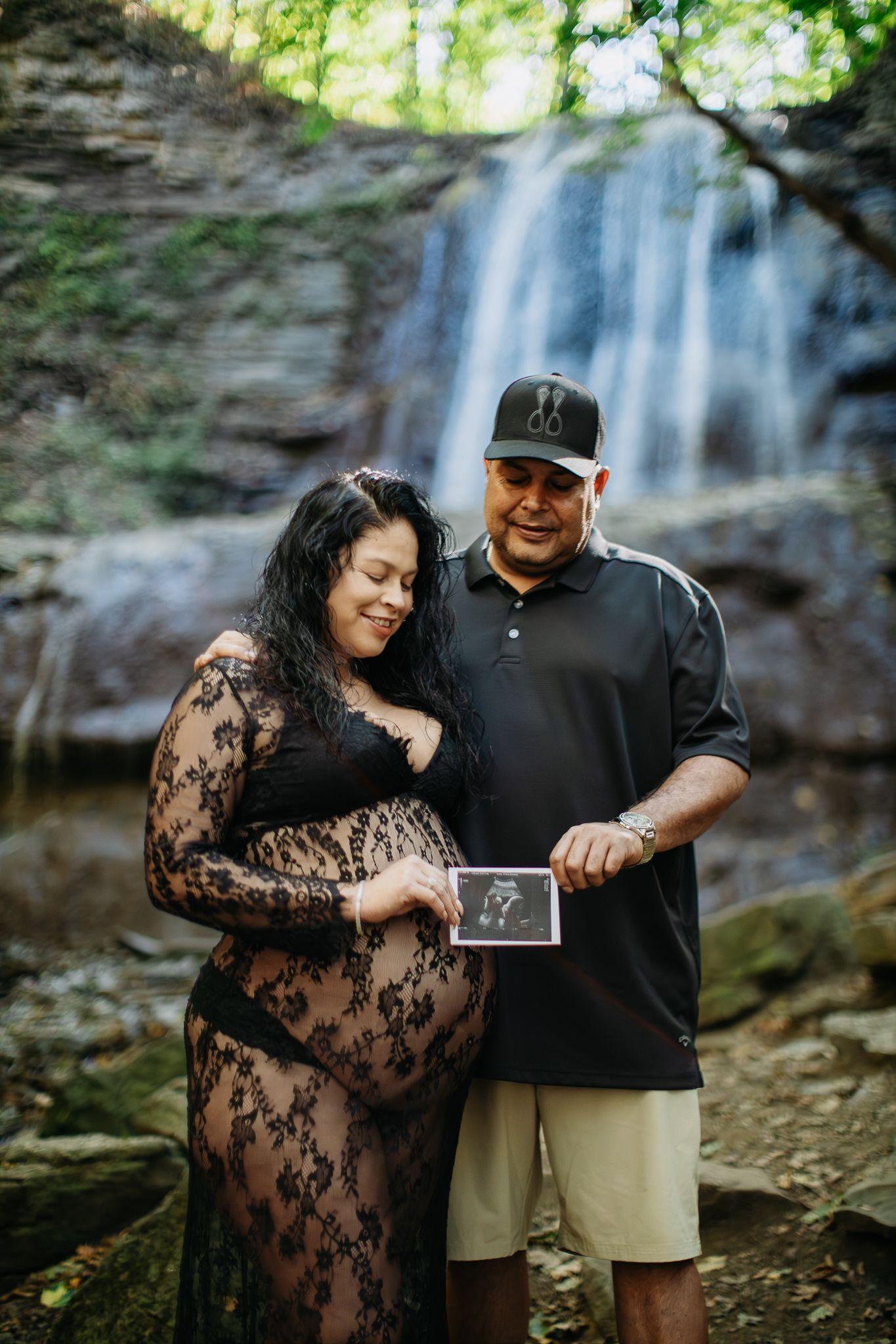 Couple sharing happy moments while displaying ultrasound photos during their waterfall photoshoot.