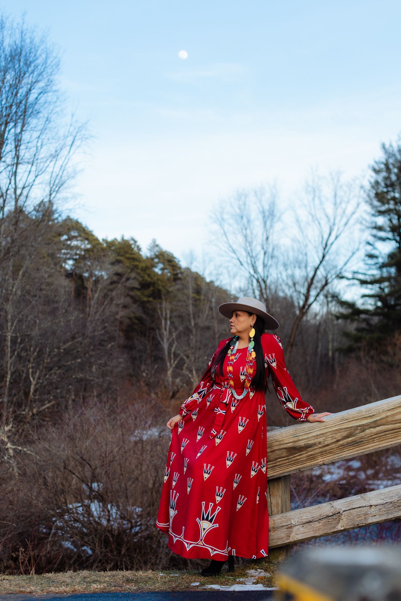 Native American woman in red traditional dress and hat leans on wooden fence at dusk with moon visible.