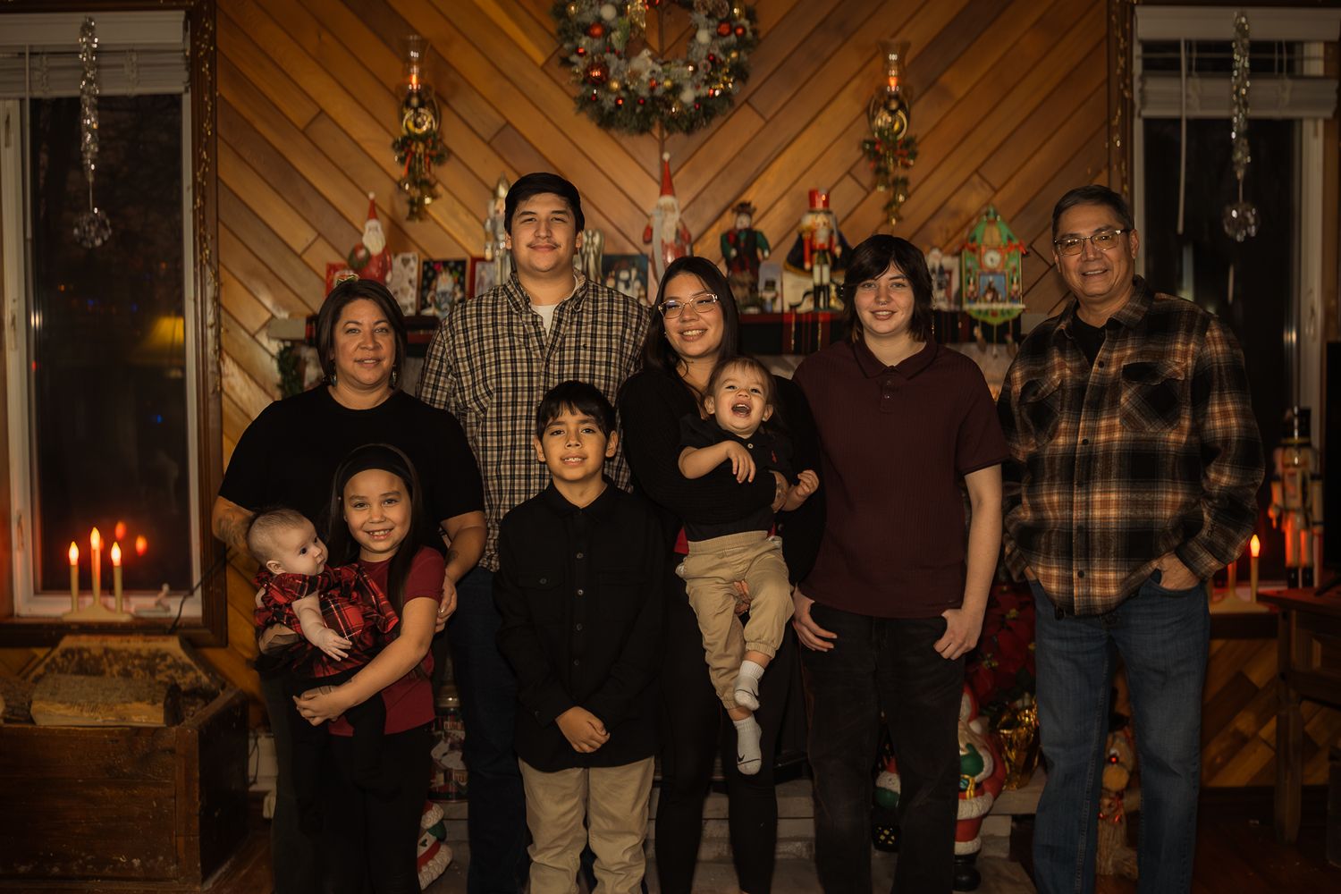 Family of eight poses together indoors for a festive Christmas portrait with holiday decorations and candlelight in background.