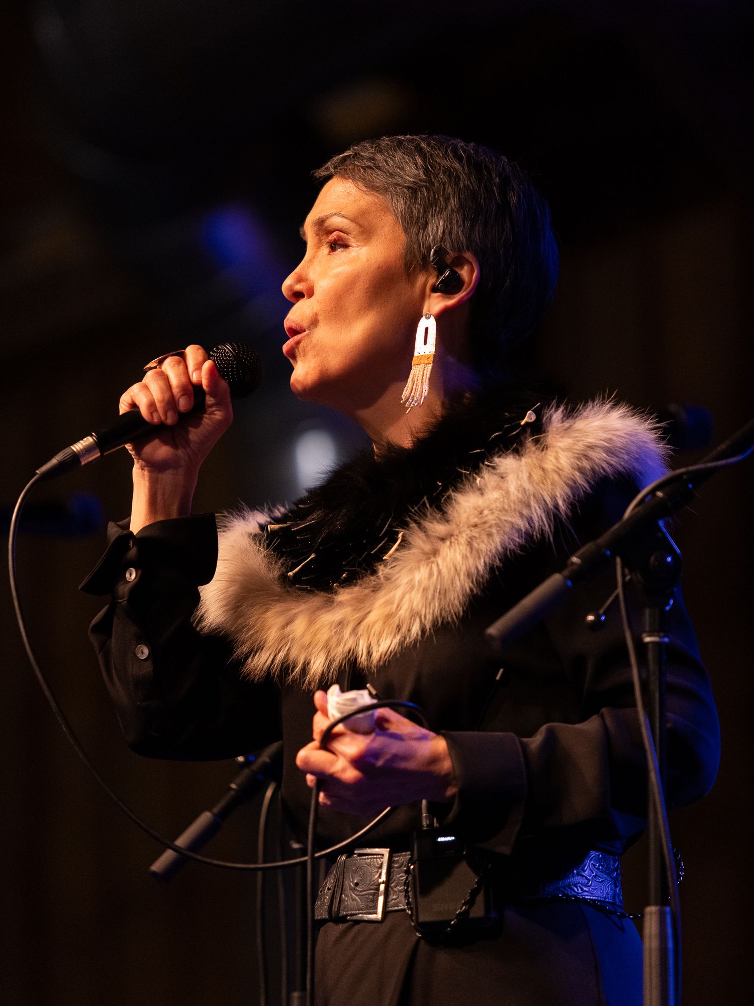 A woman with short hair sings into a microphone on stage wearing a black outfit with a fur-trimmed collar.