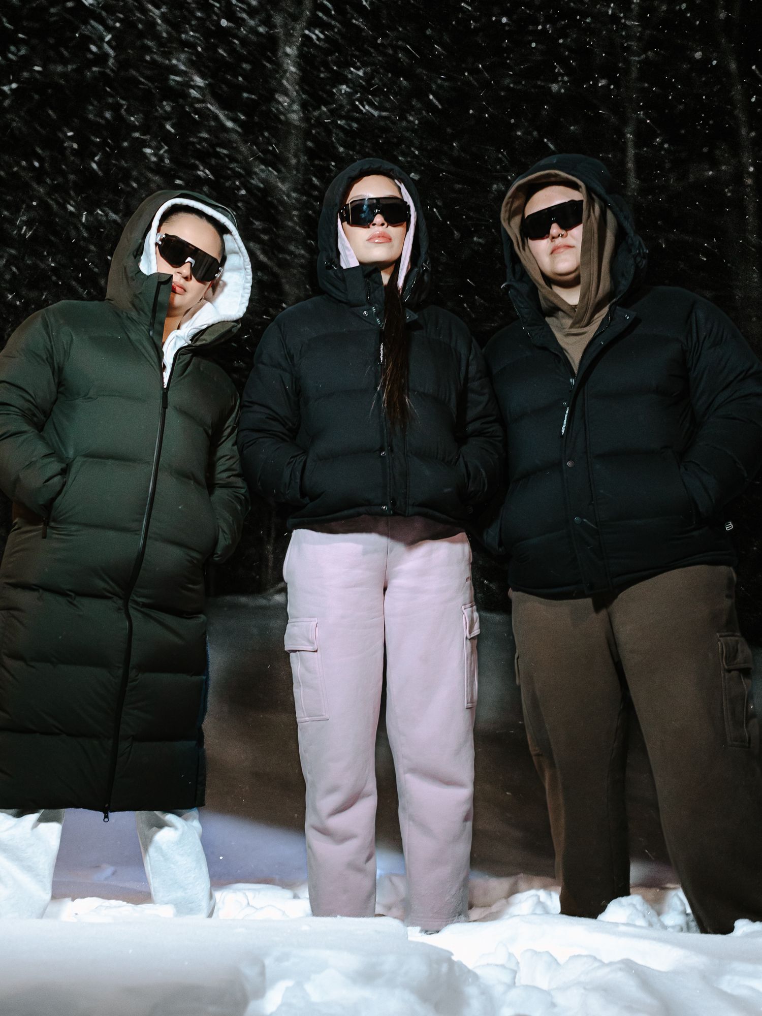 Three women in hooded puffer jackets and sunglasses pose in snowy winter setting at night.