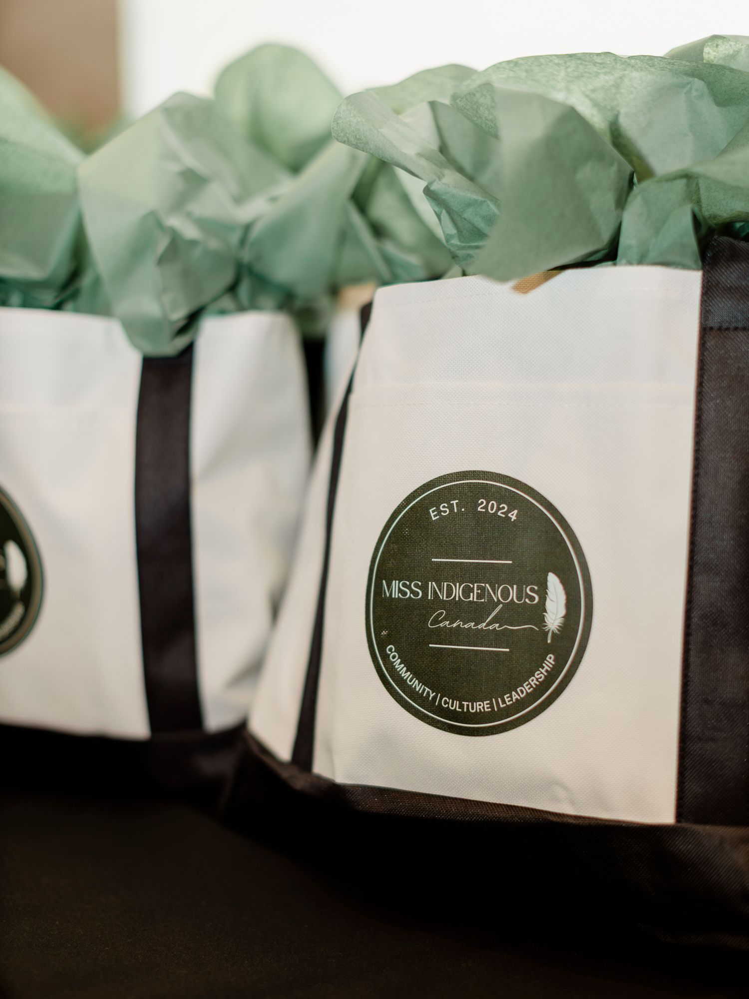 White tote bags with Miss Indigenous Canada Est. 2024 logo filled with green tissue paper at an event.