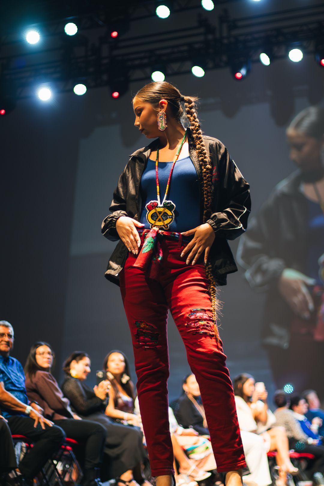 Model walks runway in red distressed jeans, blue top, black jacket, and colorful beaded necklace at fashion show.
