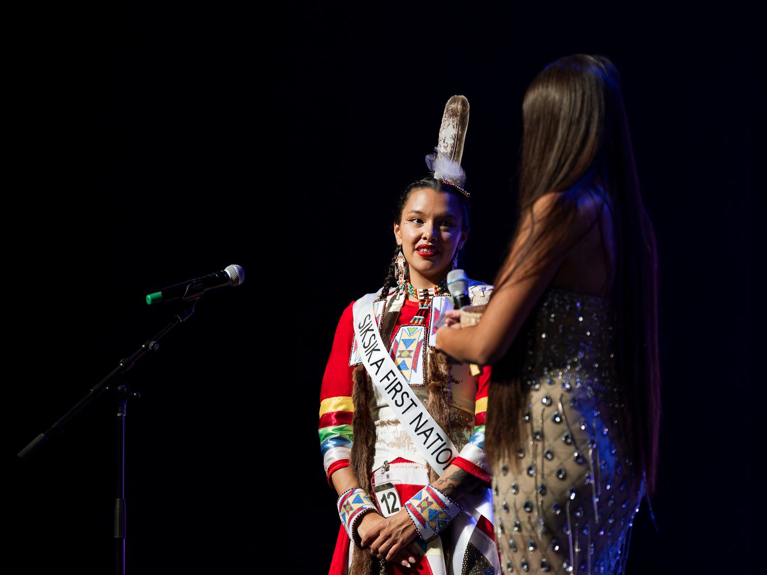 Native First Nations pageant contestant speaks into microphone on stage wearing traditional red regalia and sash.