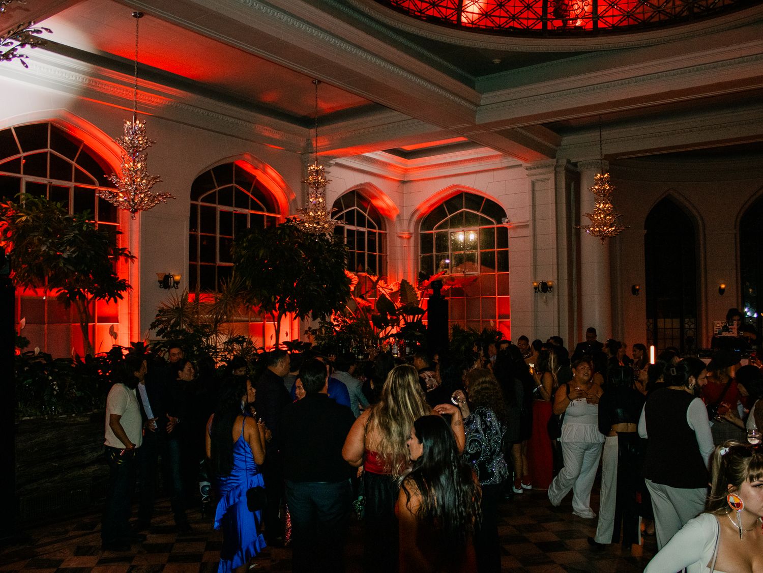 Elegant ballroom filled with guests dancing under red ambient lighting and chandeliers.