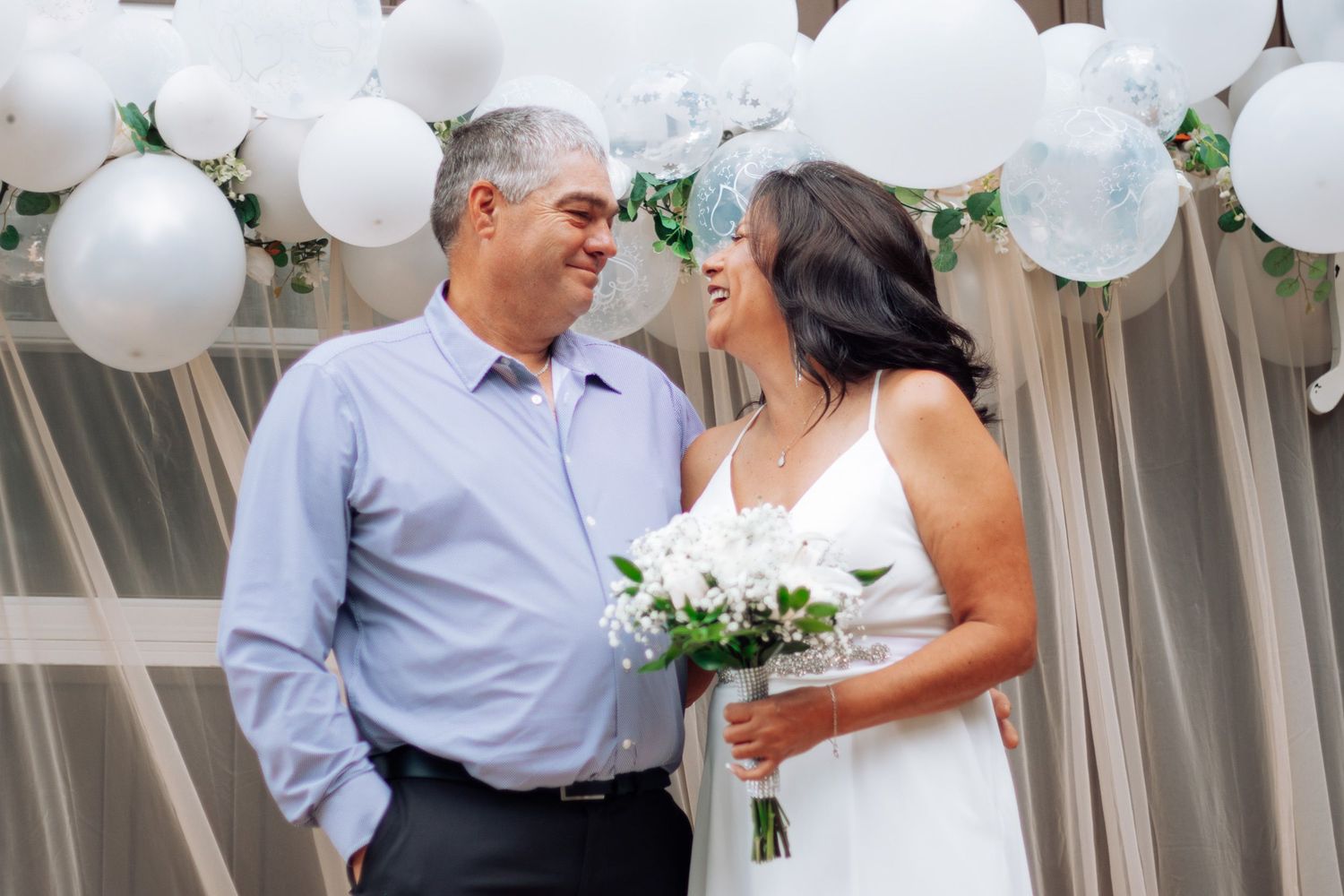 Bride in white dress holding white bouquet smiling at groom under white balloon arch at wedding ceremony.