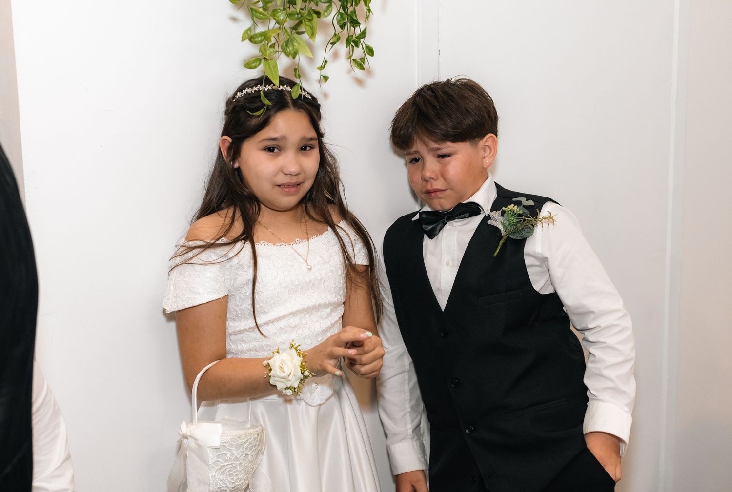 A young flower girl in white dress and boy in tuxedo pose together at a wedding ceremony.