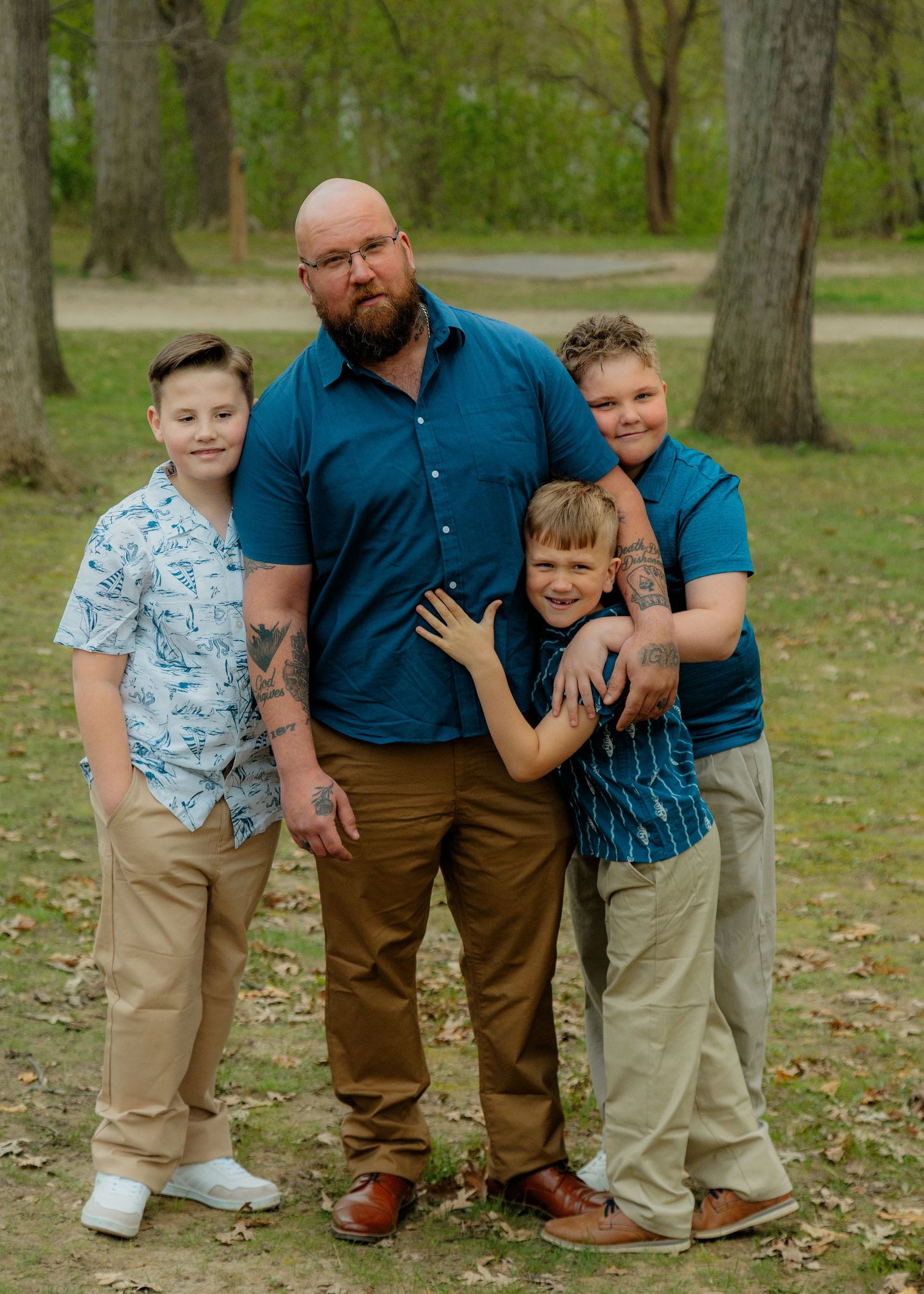 A man and three boys pose together outdoors in a park setting, dressed in coordinating blue and khaki outfits.