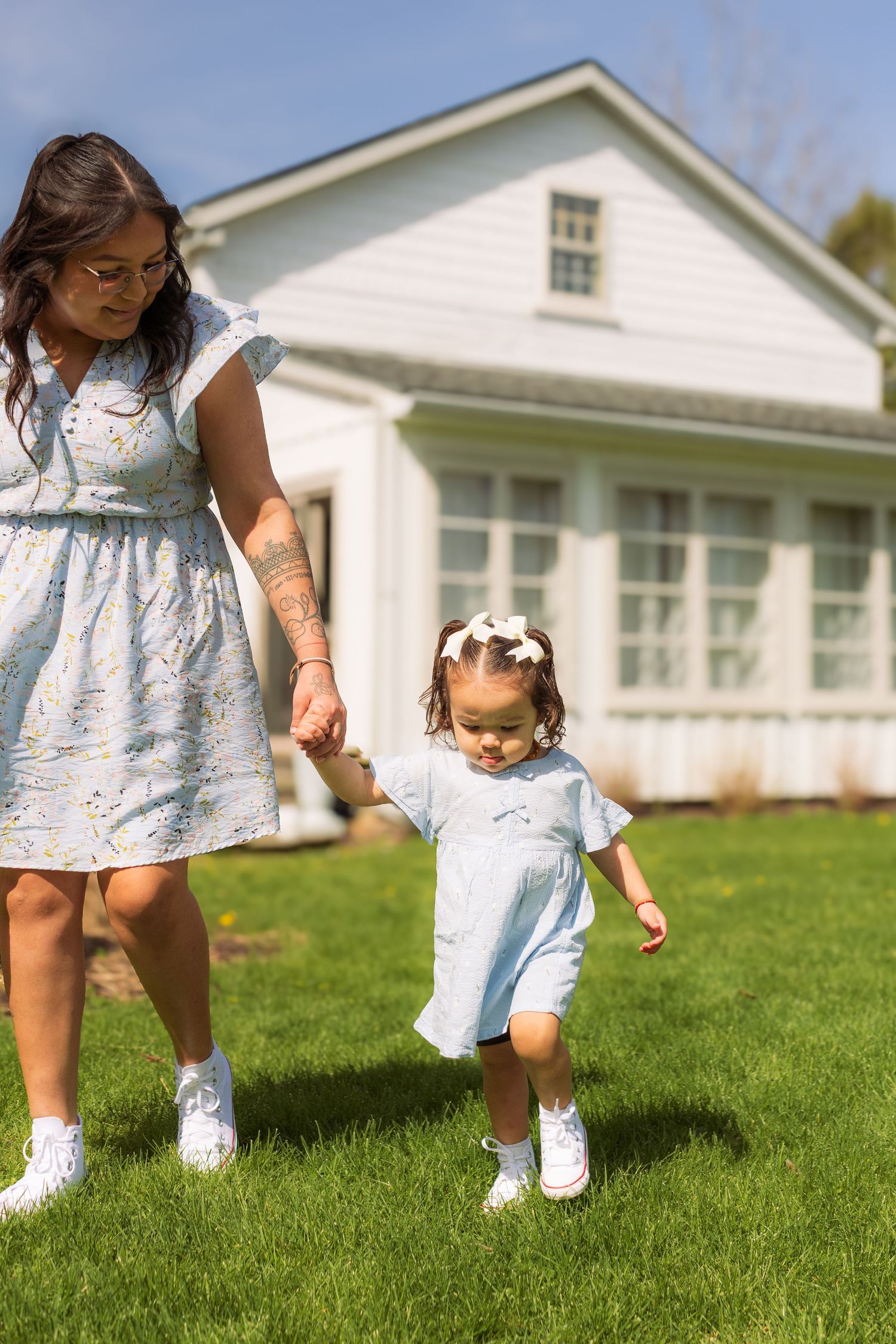 A mother and toddler in matching light blue outfits hold hands while walking on green grass in front of a white house.