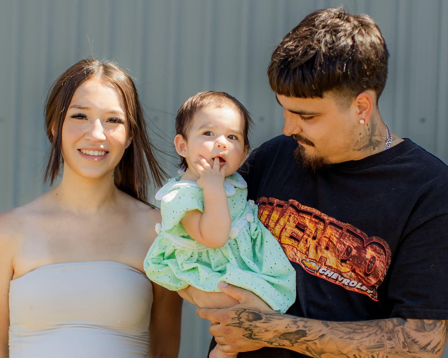 Young parents smile holding their baby girl dressed in green at an outdoor birthday celebration.