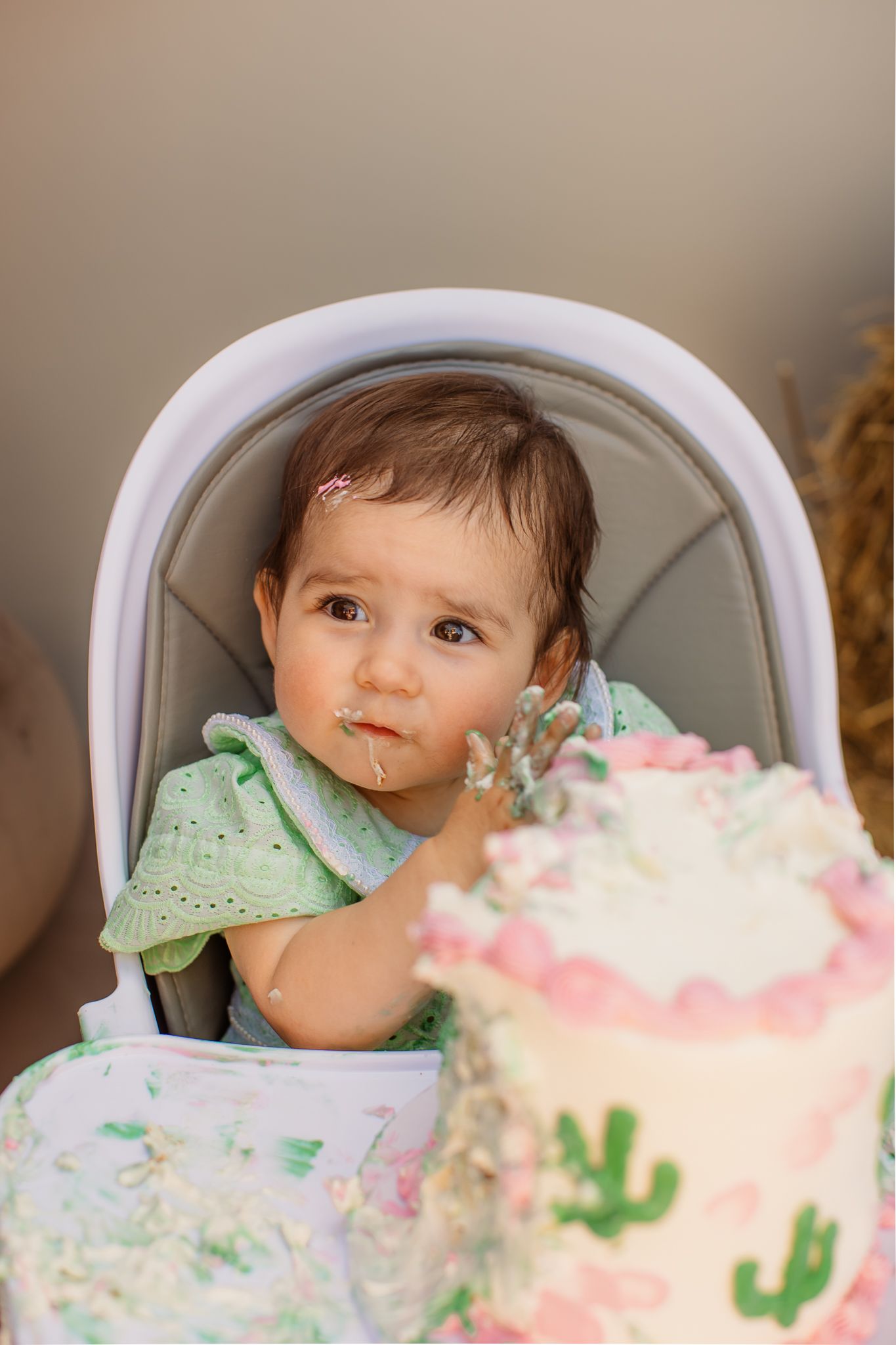 Baby girl in green outfit smashing first birthday cake decorated with cactus and pink flowers in high chair.