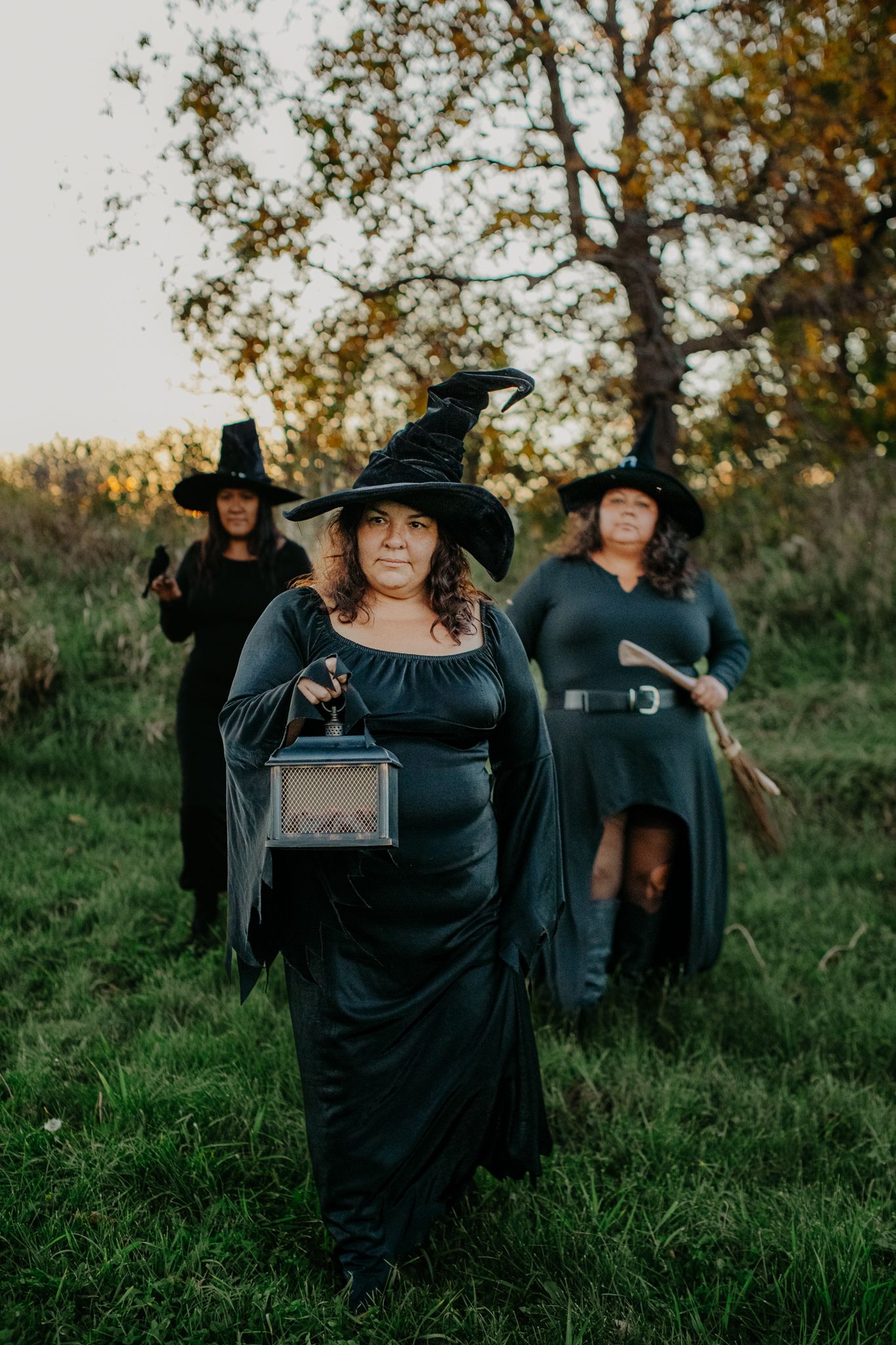 Witches in black dresses and hats posing dramatically in autumn field setting.