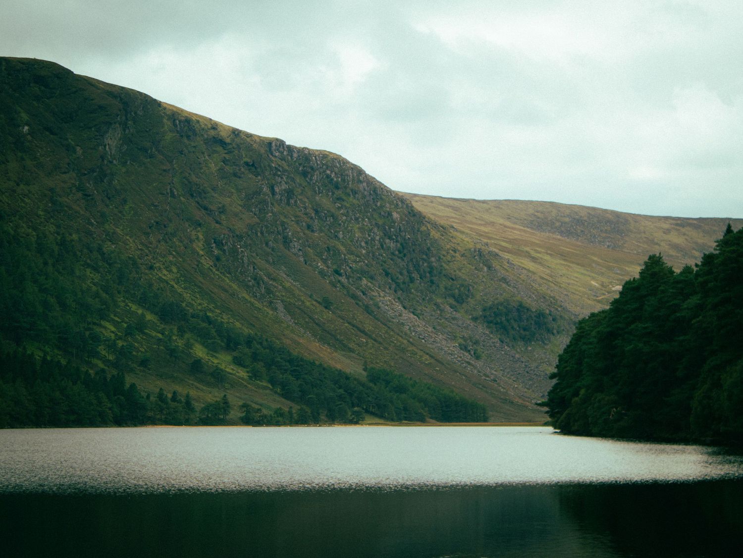 A moody landscape of a lake surrounded by mountains under an overcast sky.