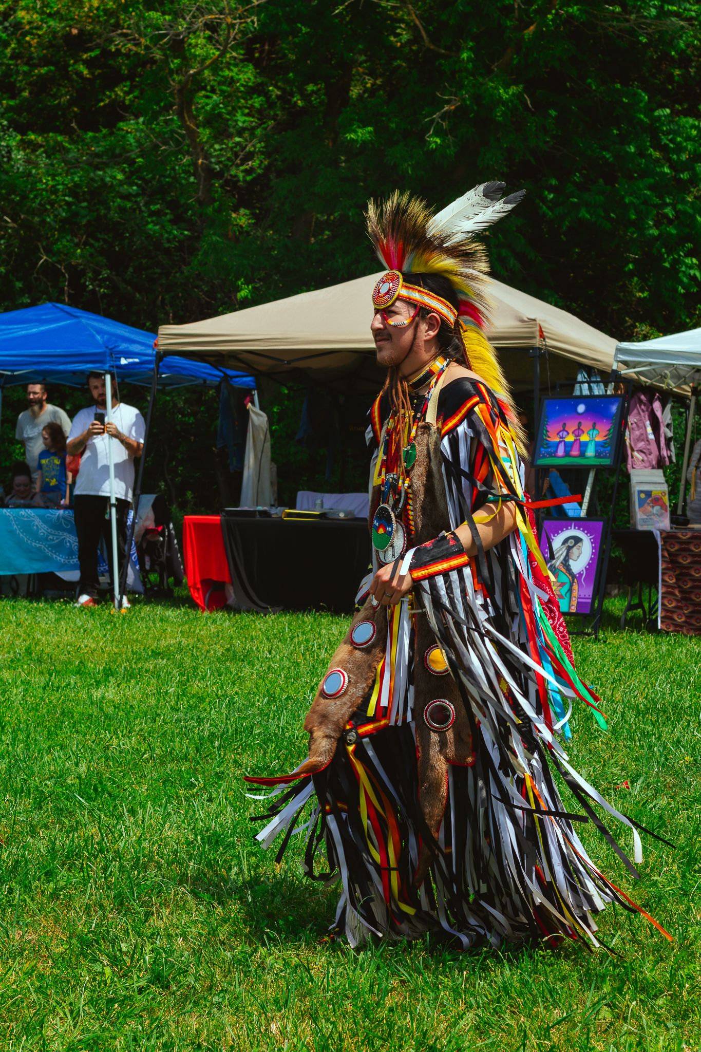 Traditional Native American dancers in regalia performing on grass field near vendor tents.