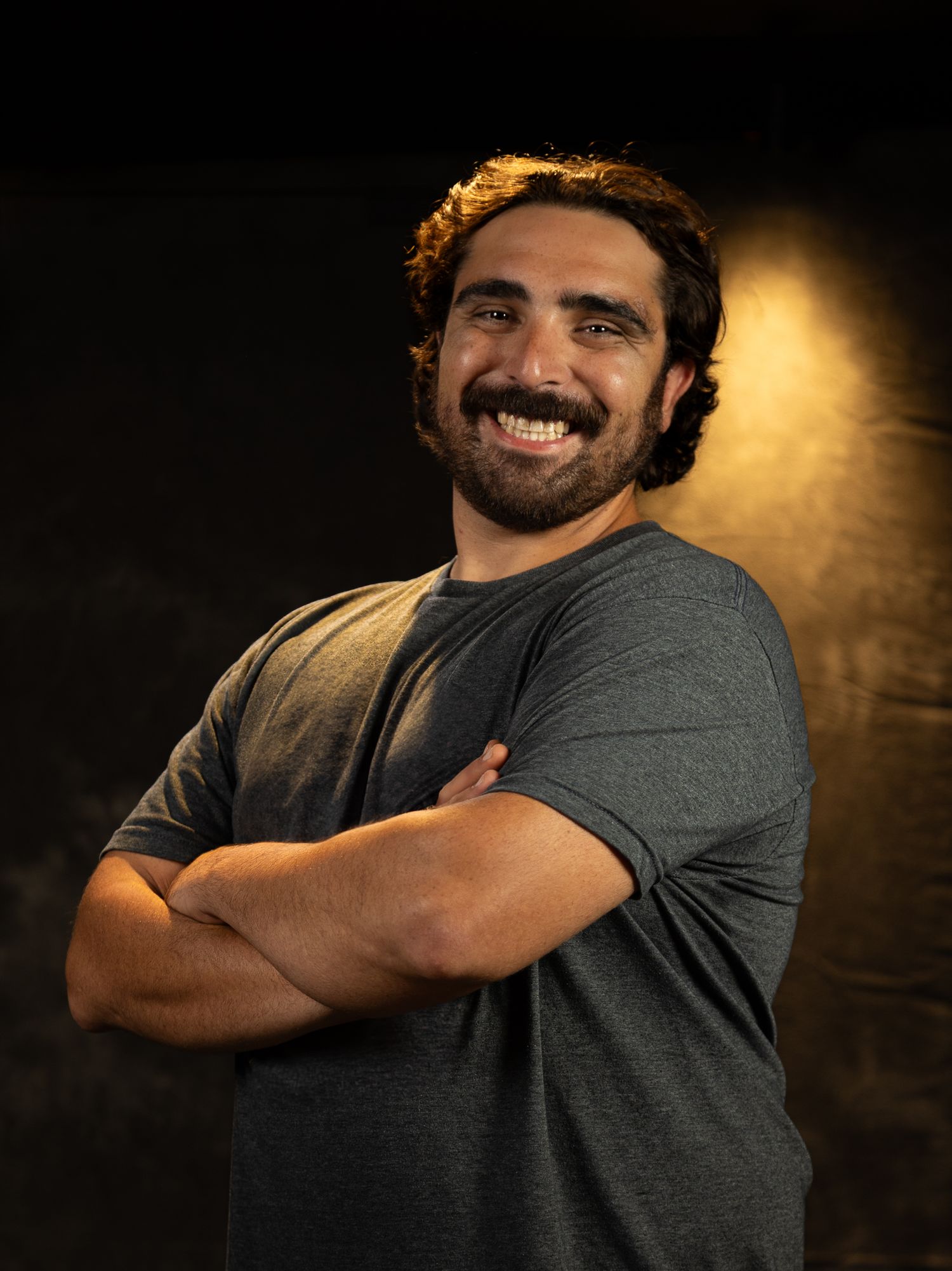 Studio portrait of person in grey shirt with crossed arms against moody background.